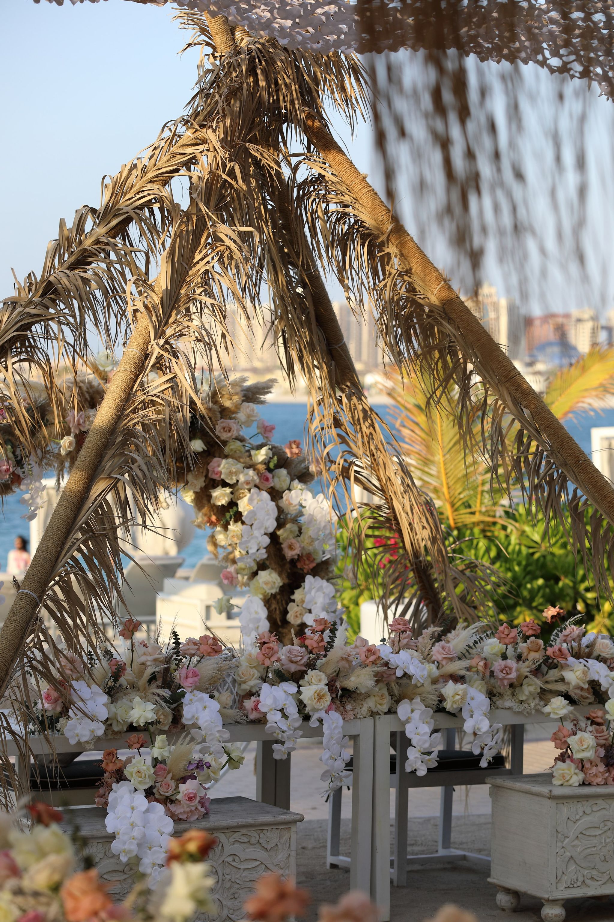 Dried palm-frond arch with cascading blush and ivory flowers, the sea and city towers beyond