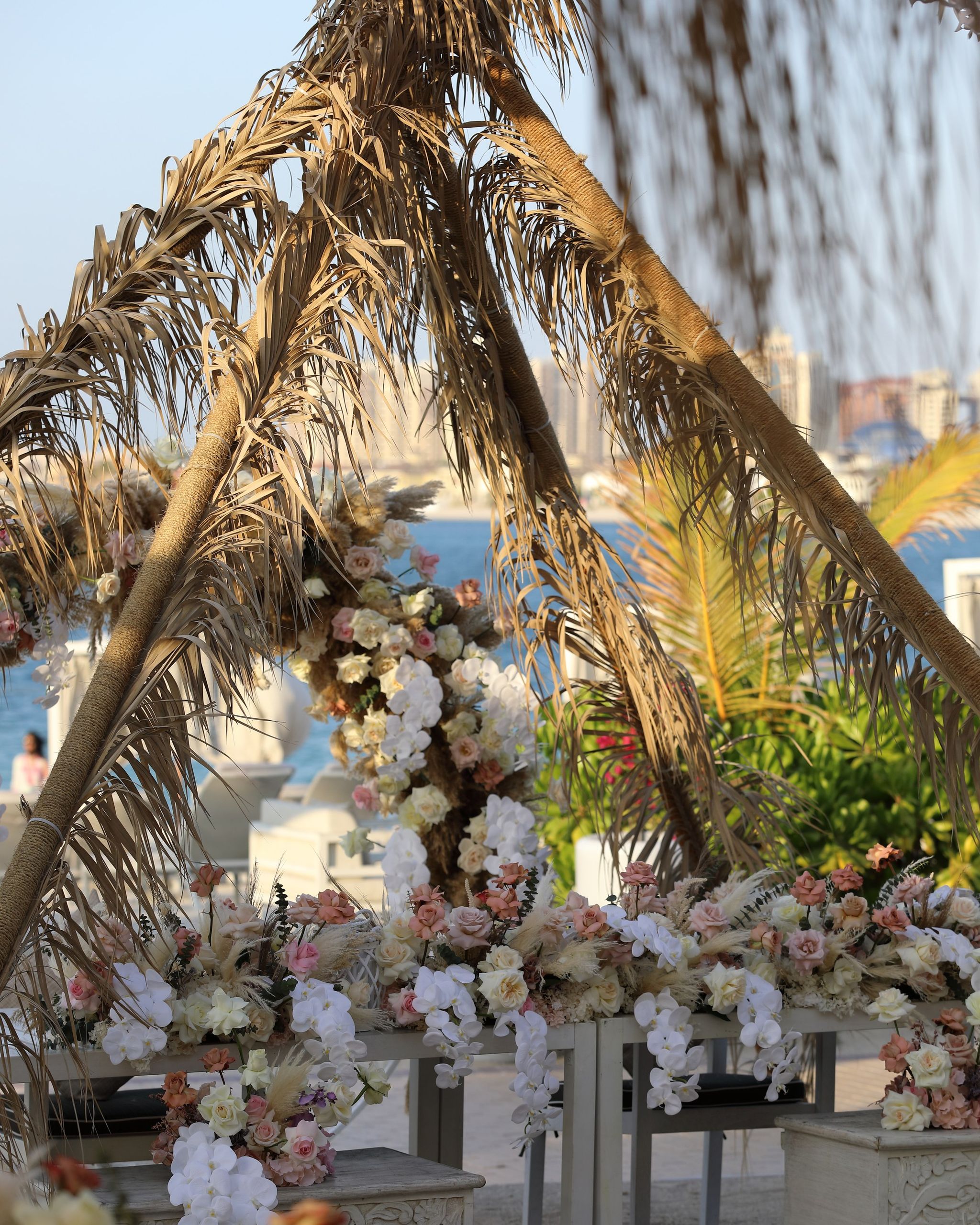 Dried palm-frond arch with cascading blush and ivory flowers, the sea and city towers beyond