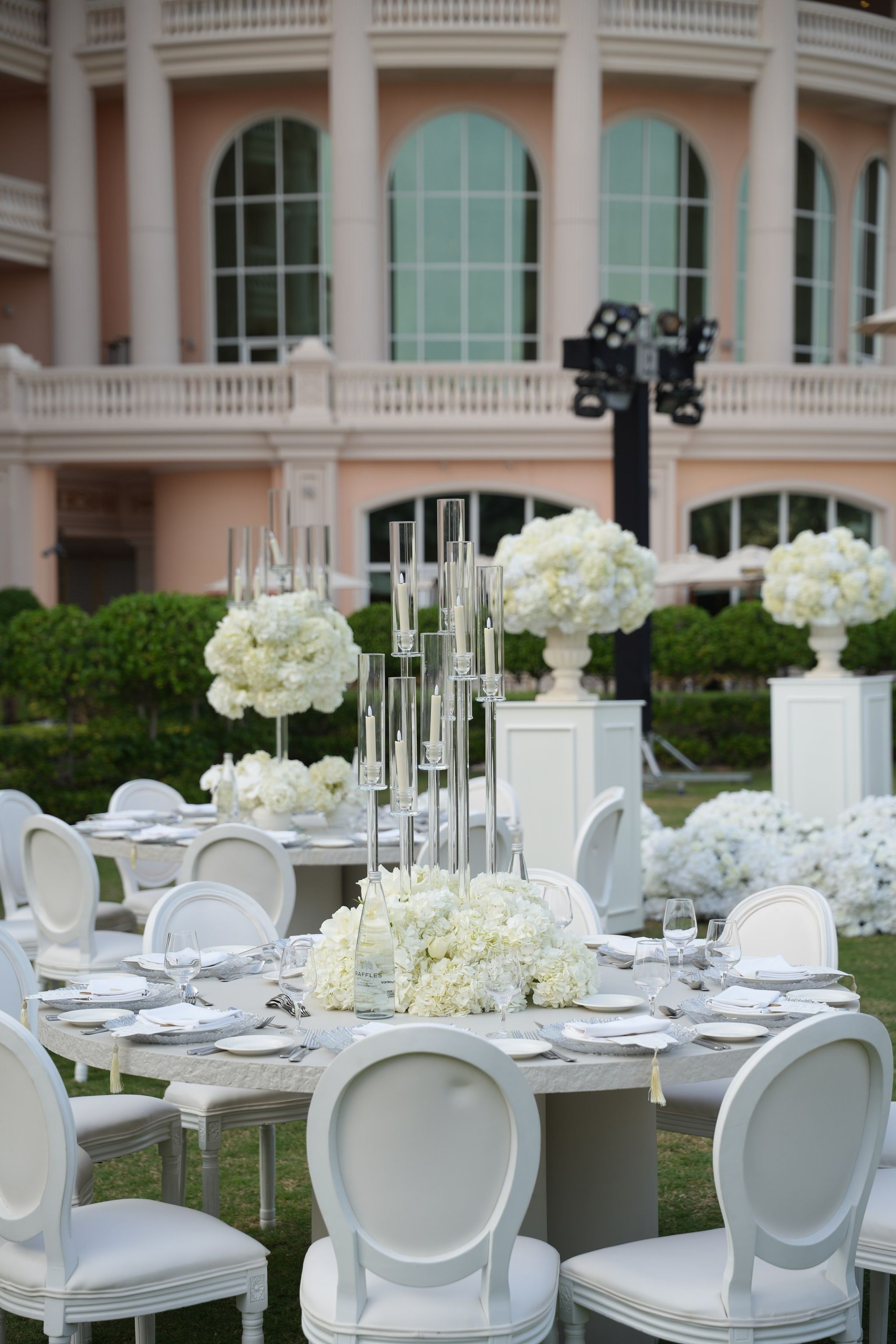 Reception tables with crystal candelabra and white hydrangeas on the lawn before the arched windows of Raffles The Palm Dubai