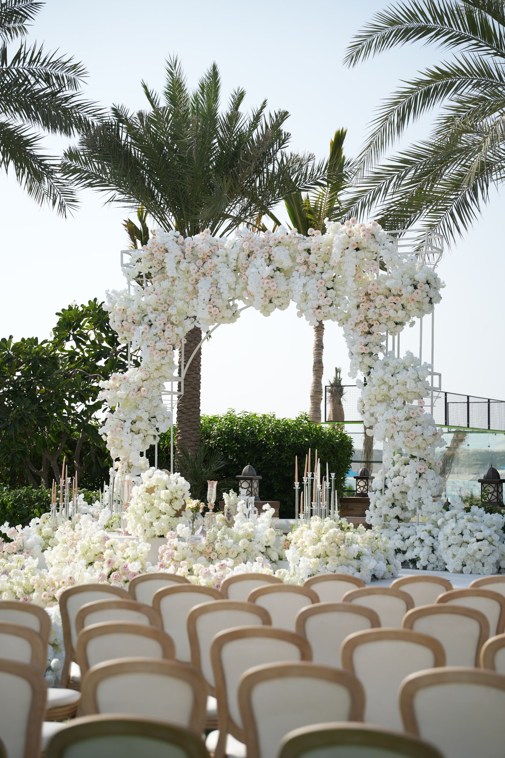 White and blush rose arch rising above rows of ivory ceremony chairs, framed by date palms