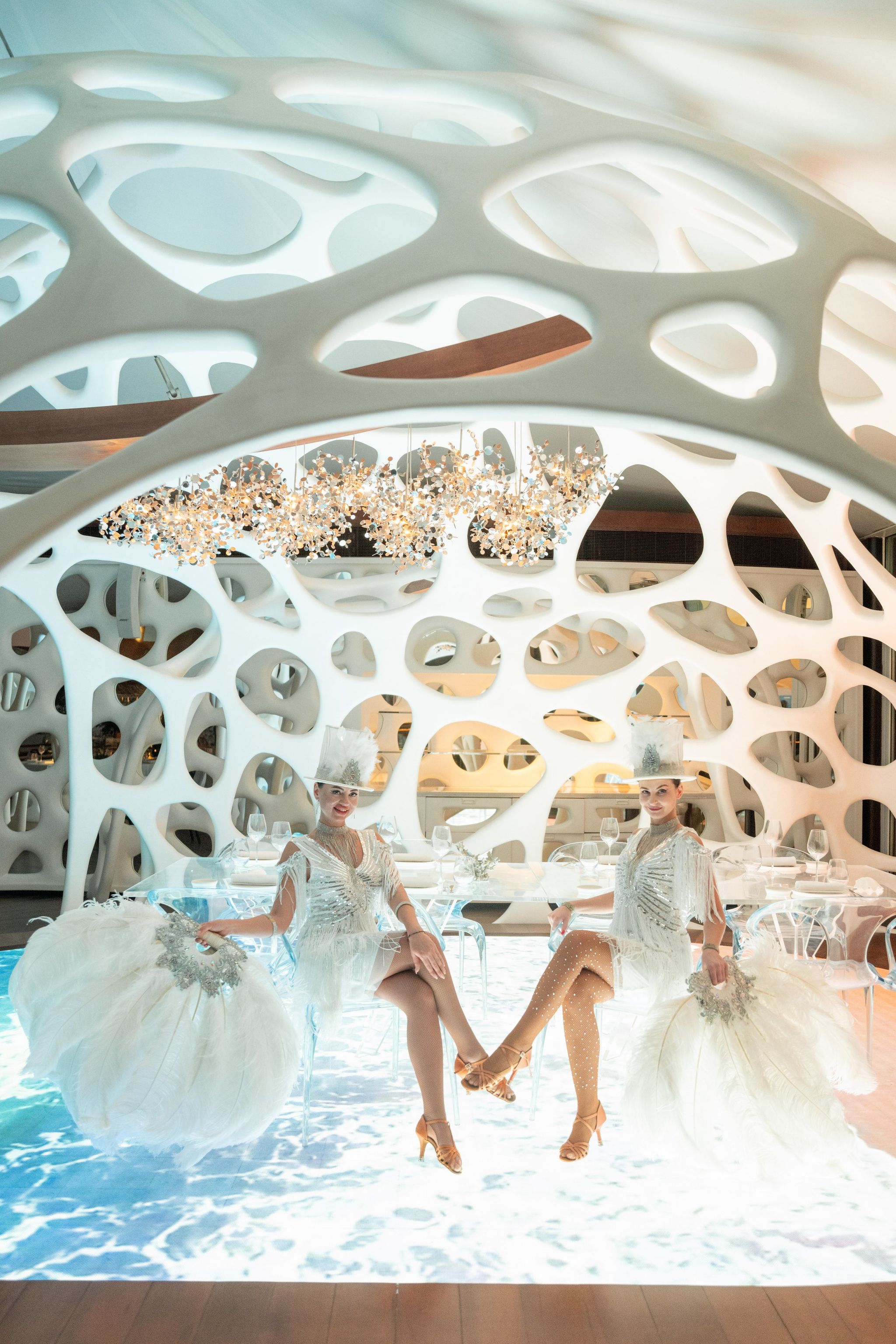 Two feathered performers seated beneath a sculpted white arch above a projected sea floor
