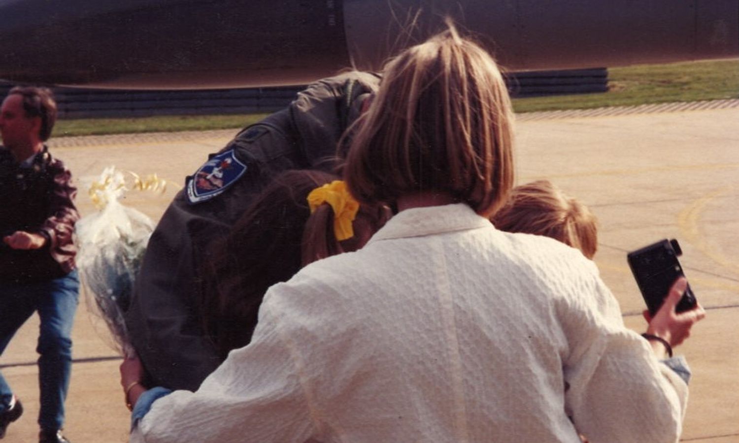 Spring 1991 — flight suit, flowers, a Polaroid in someone's hand, and three small arms holding on.
