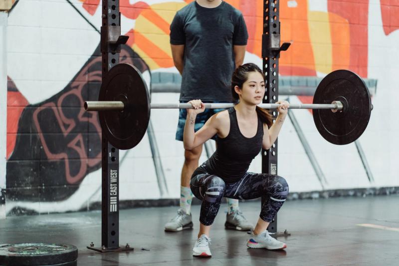 A woman squats with a loaded barbell across her shoulders in a squat rack while a spotter stands behind her.