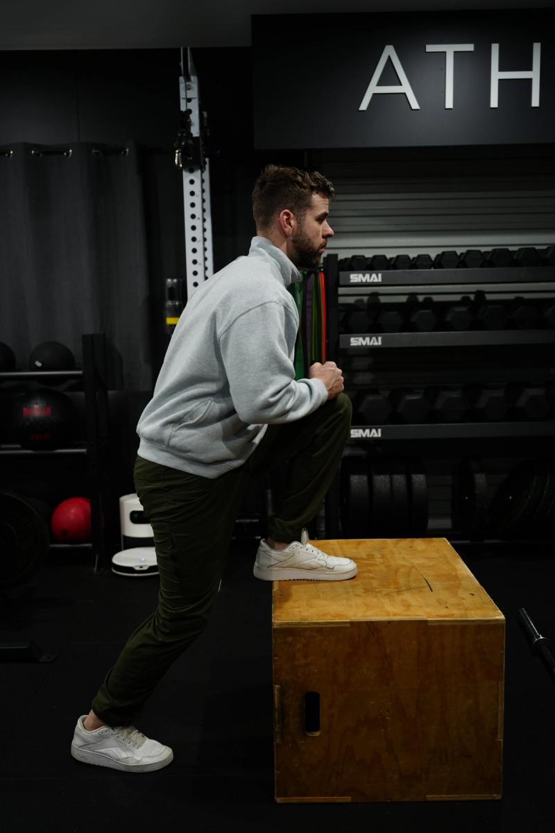 A man steps one foot up onto a wooden plyo box during a step-up exercise in a gym.