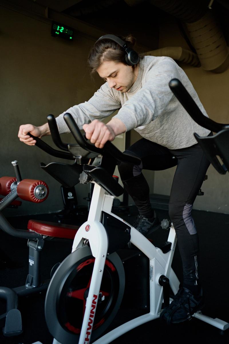 A man in headphones and athletic wear leans forward riding a white Schwinn indoor cycling bike in a gym.