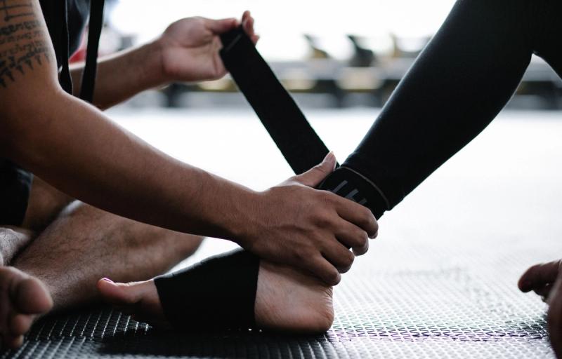 Hands wrap a black support strap around a person's ankle and foot on a black gym mat.