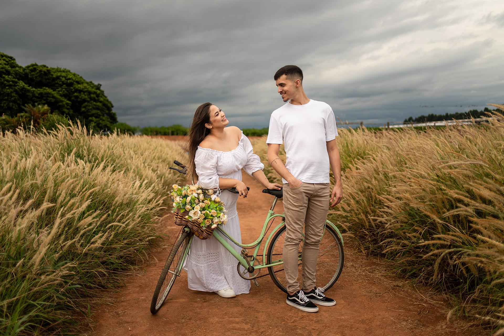 Casal em uma estrada de terra entre capim dourado, ela segurando uma bicicleta com flores
