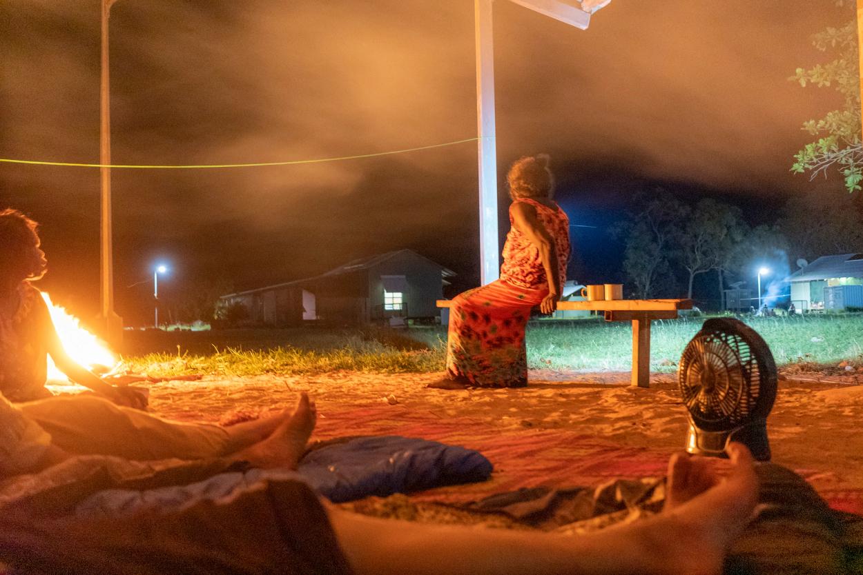 Night time at a campsite, woman sits facing away from a fire atop the sand