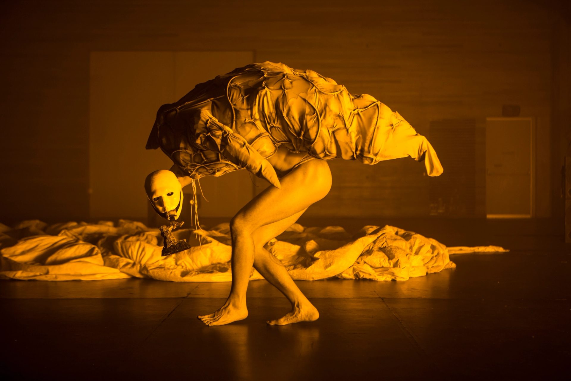 Performance art where legs are bare and head (a white mask) is extended in front of a slightly folded body that carries a kind of chrysalis.