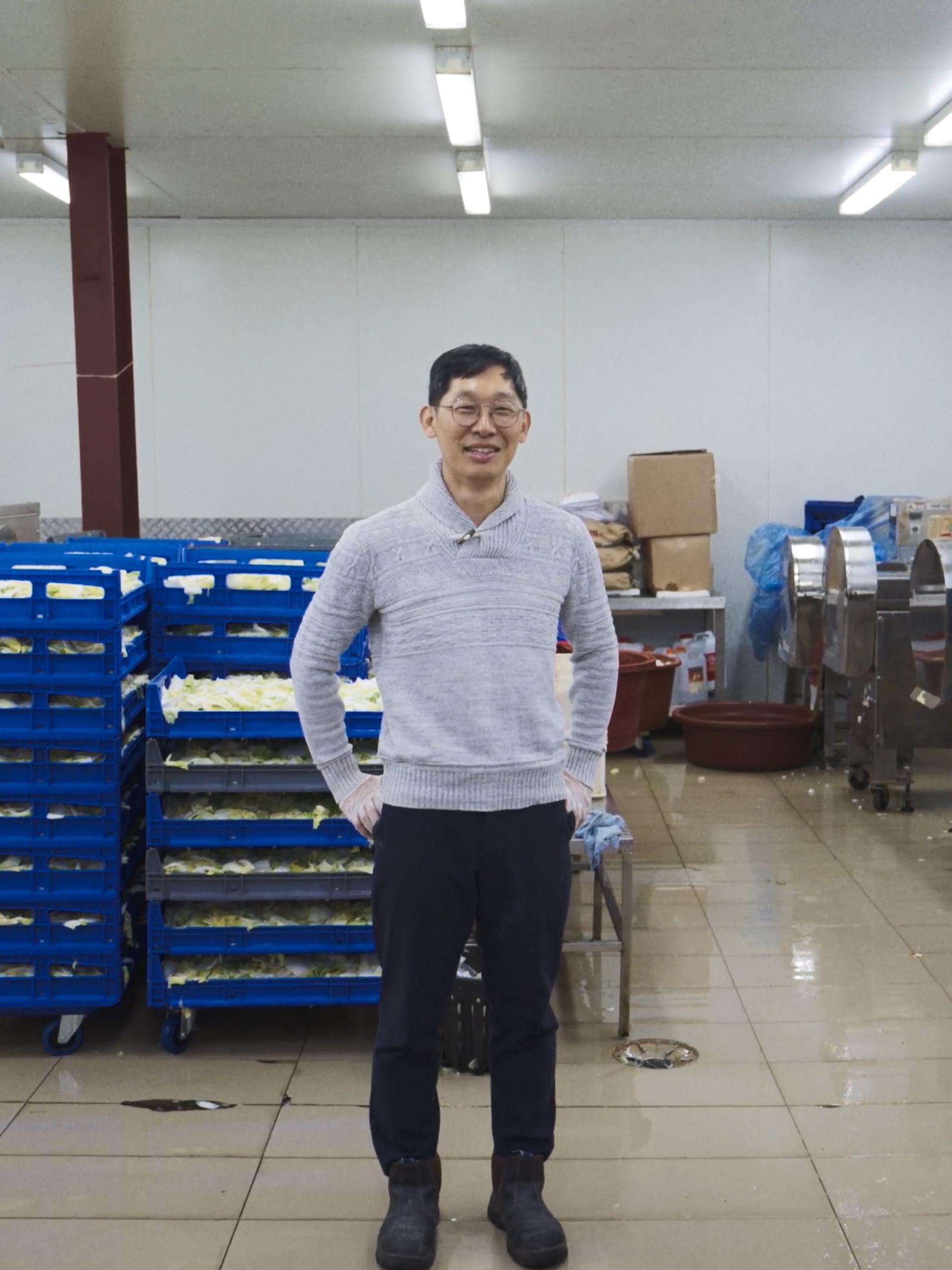 A man standing in an indoor manufacturing factory, with pallets of cabbage behind him.
