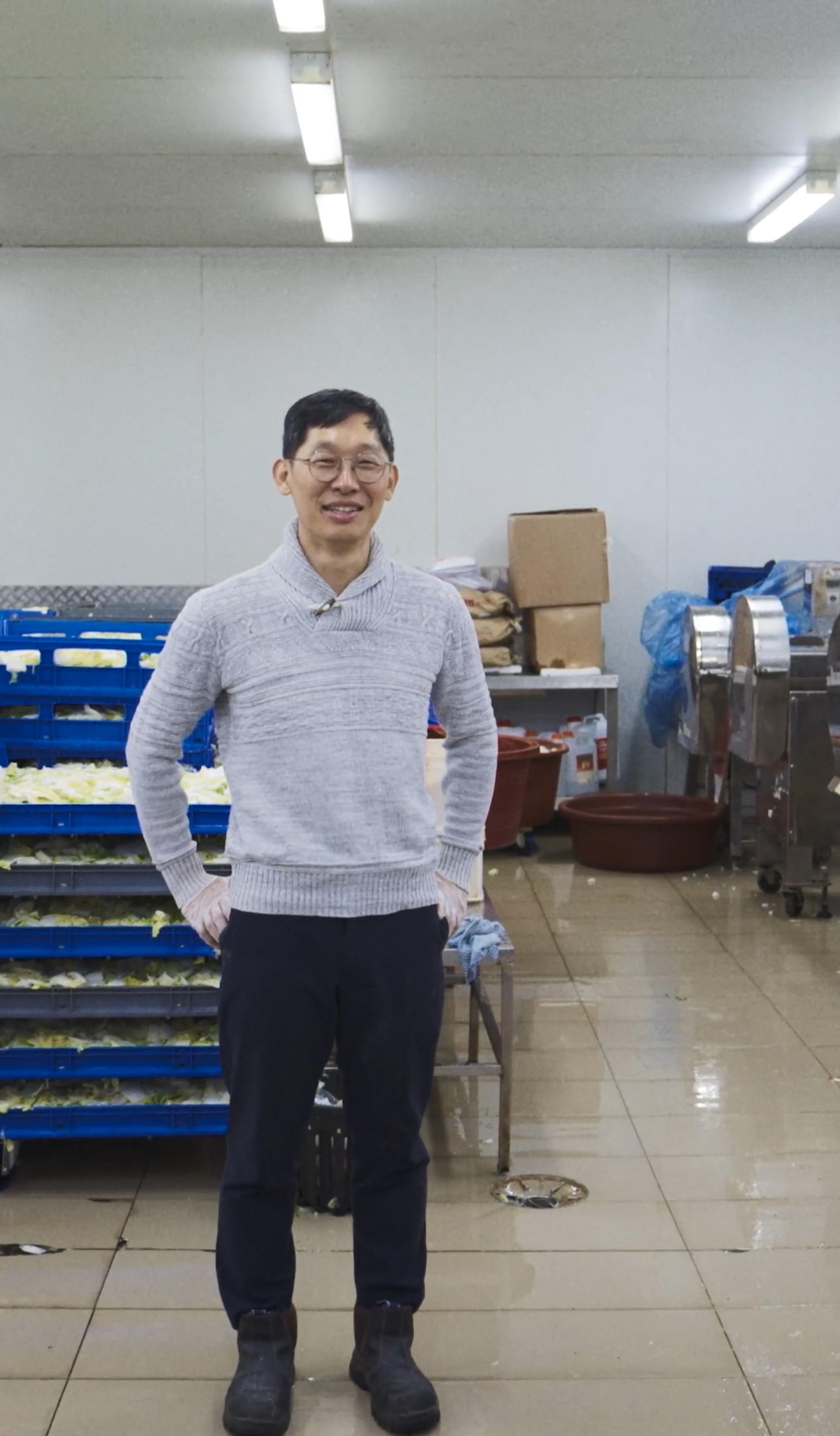 A man standing in an indoor manufacturing factory, with pallets of cabbage behind him.