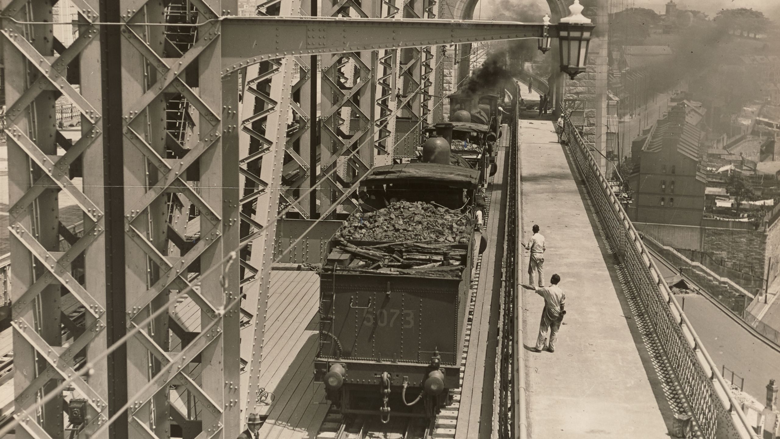 Landscape format photographic image depicting a stage in the construction of the Sydney Harbour Bridge. Looking south at trains with tenders used for load tests on the outer west track. In th background we can see the Sydney Observatory and houses at Millers Point.