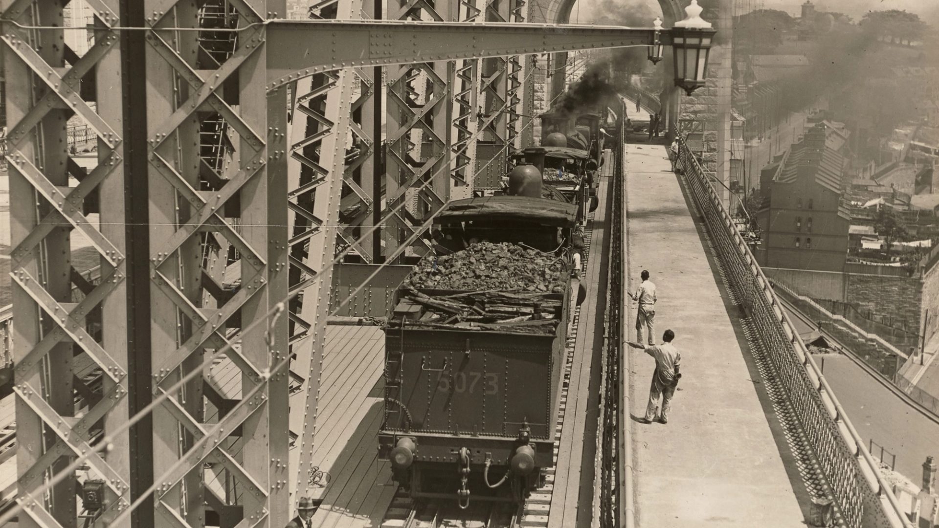Landscape format photographic image depicting a stage in the construction of the Sydney Harbour Bridge. Looking south at trains with tenders used for load tests on the outer west track. In th background we can see the Sydney Observatory and houses at Millers Point.