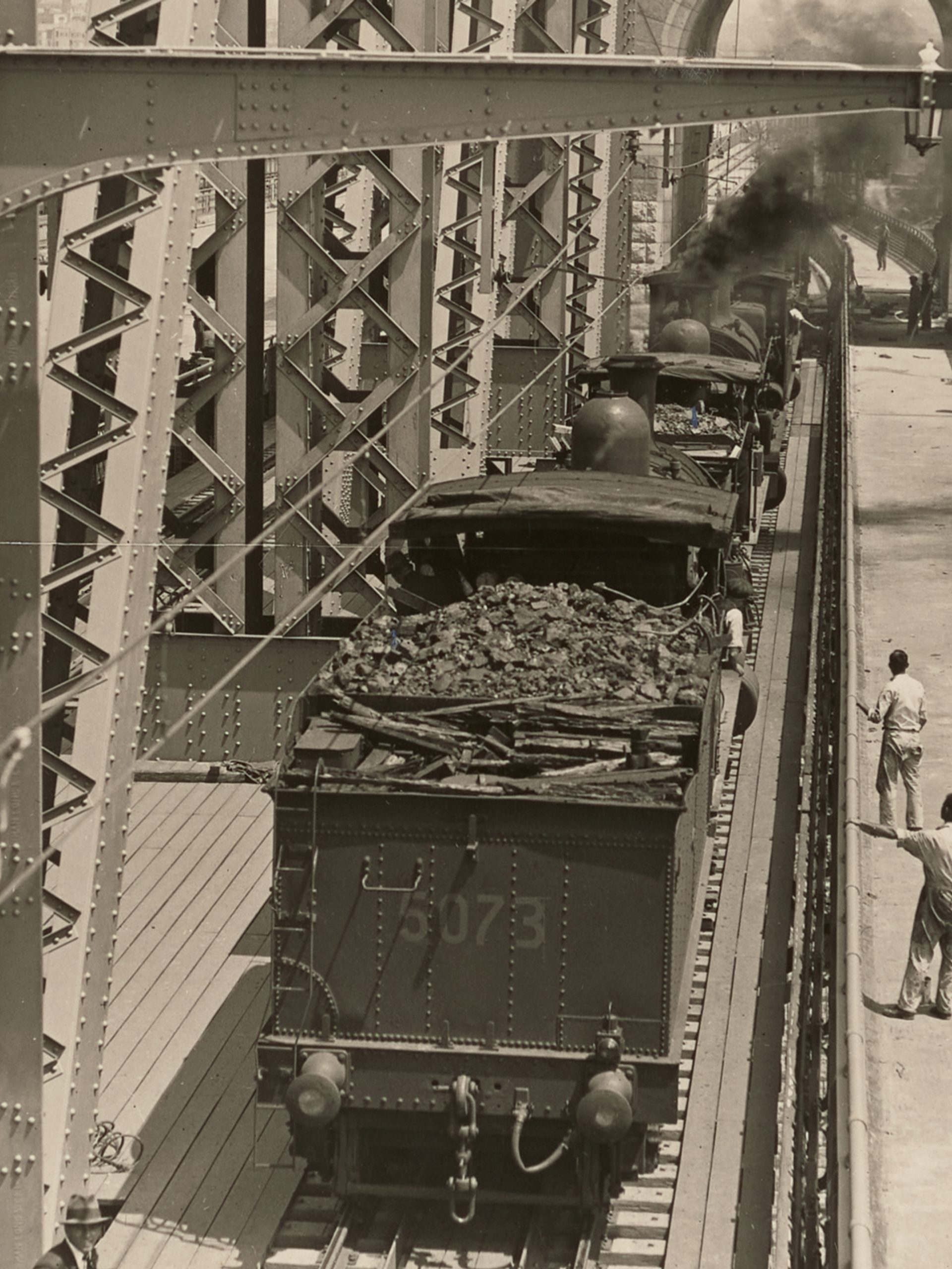 Landscape format photographic image depicting a stage in the construction of the Sydney Harbour Bridge. Looking south at trains with tenders used for load tests on the outer west track. In th background we can see the Sydney Observatory and houses at Millers Point.