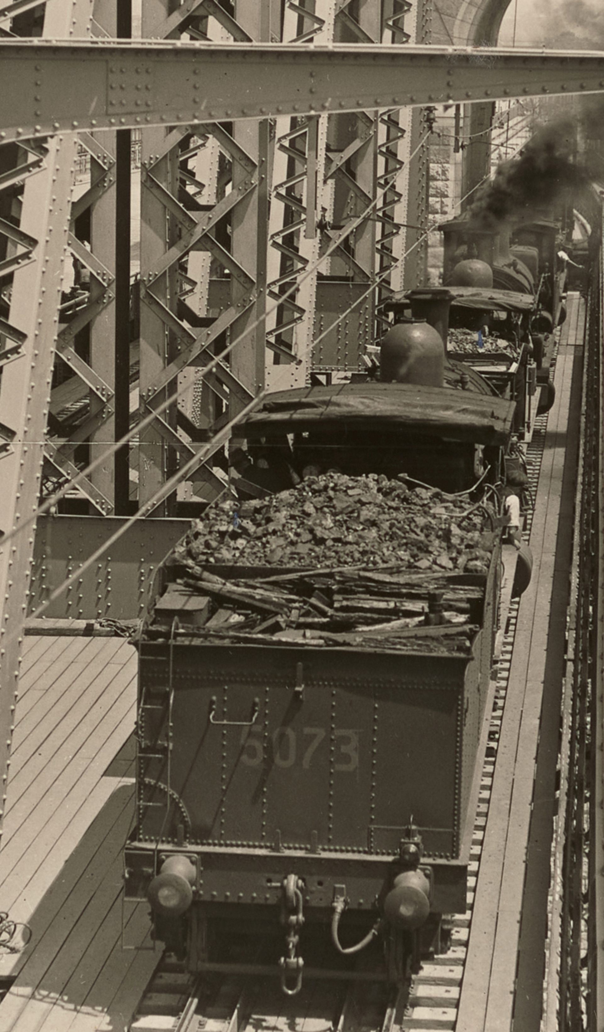 Landscape format photographic image depicting a stage in the construction of the Sydney Harbour Bridge. Looking south at trains with tenders used for load tests on the outer west track. In th background we can see the Sydney Observatory and houses at Millers Point.