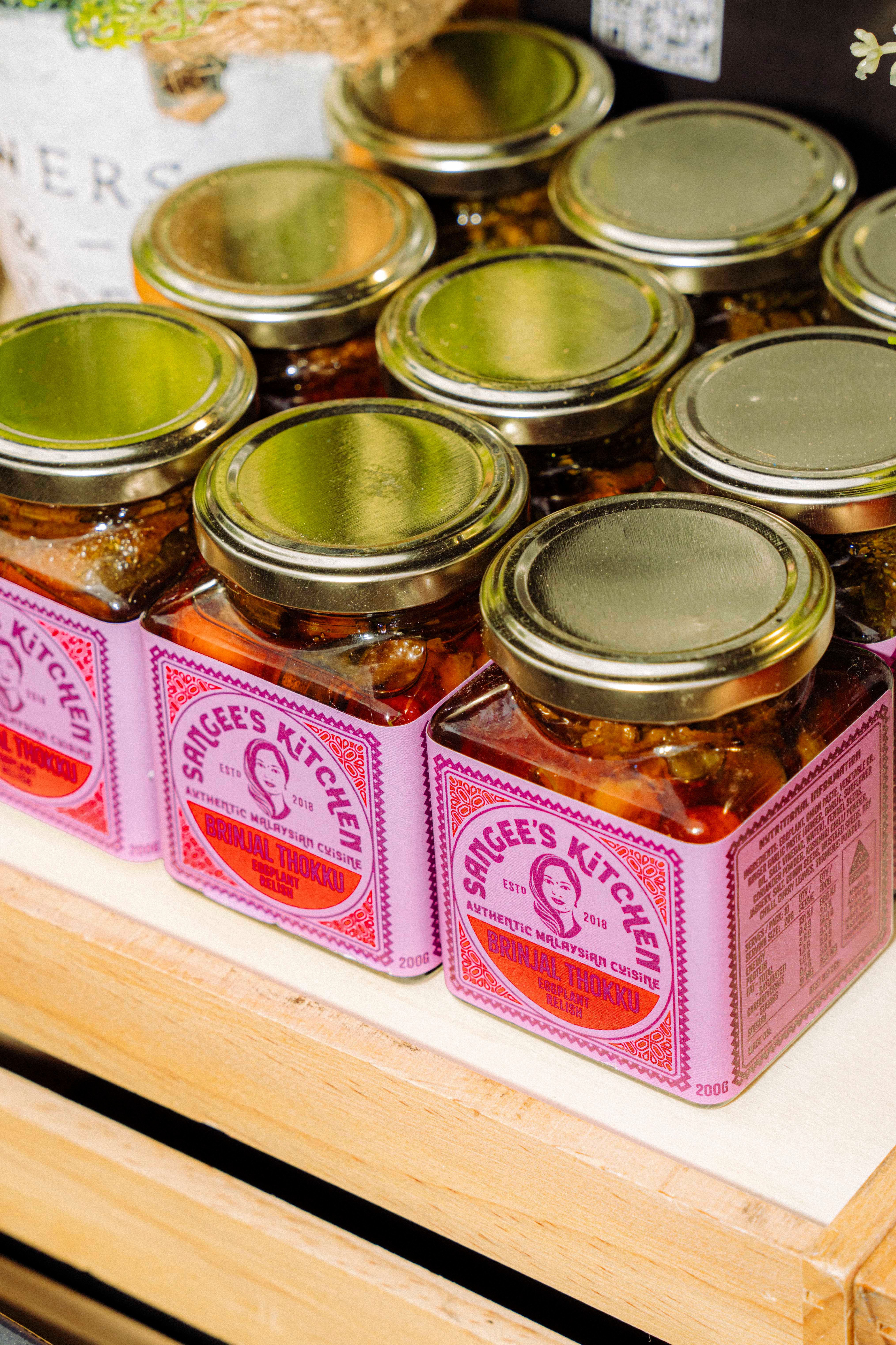 A display of jars labelled ‘Sangee's Kitchen’, containing colourful preserves or sauces, with golden lids, arranged neatly on a wooden surface.