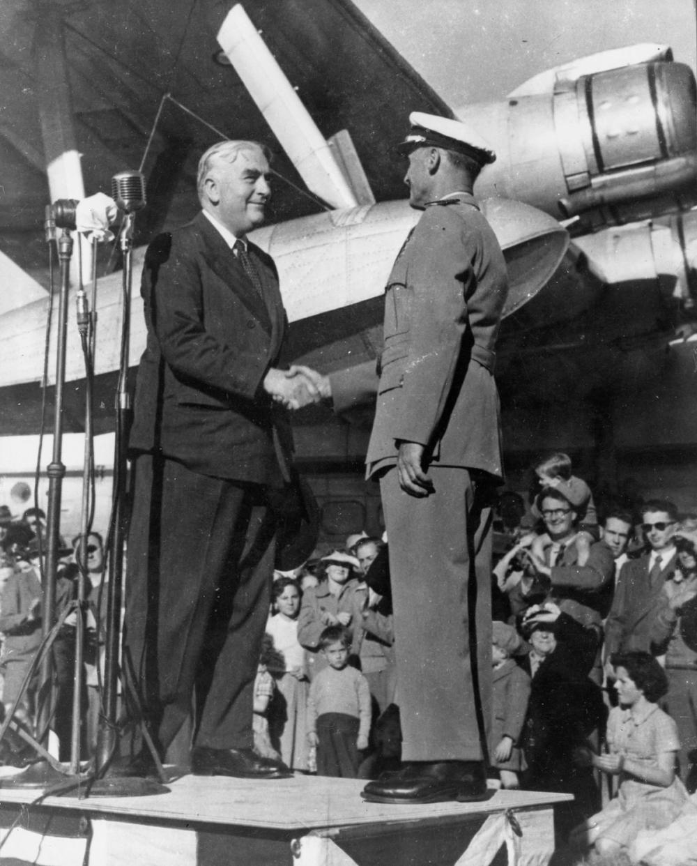 A black and white photograph of a man in a suit and a man in a pilot’s uniform standing on a podium and shaking hands. There is an audience of people behind them. Silver plane visible in the background.