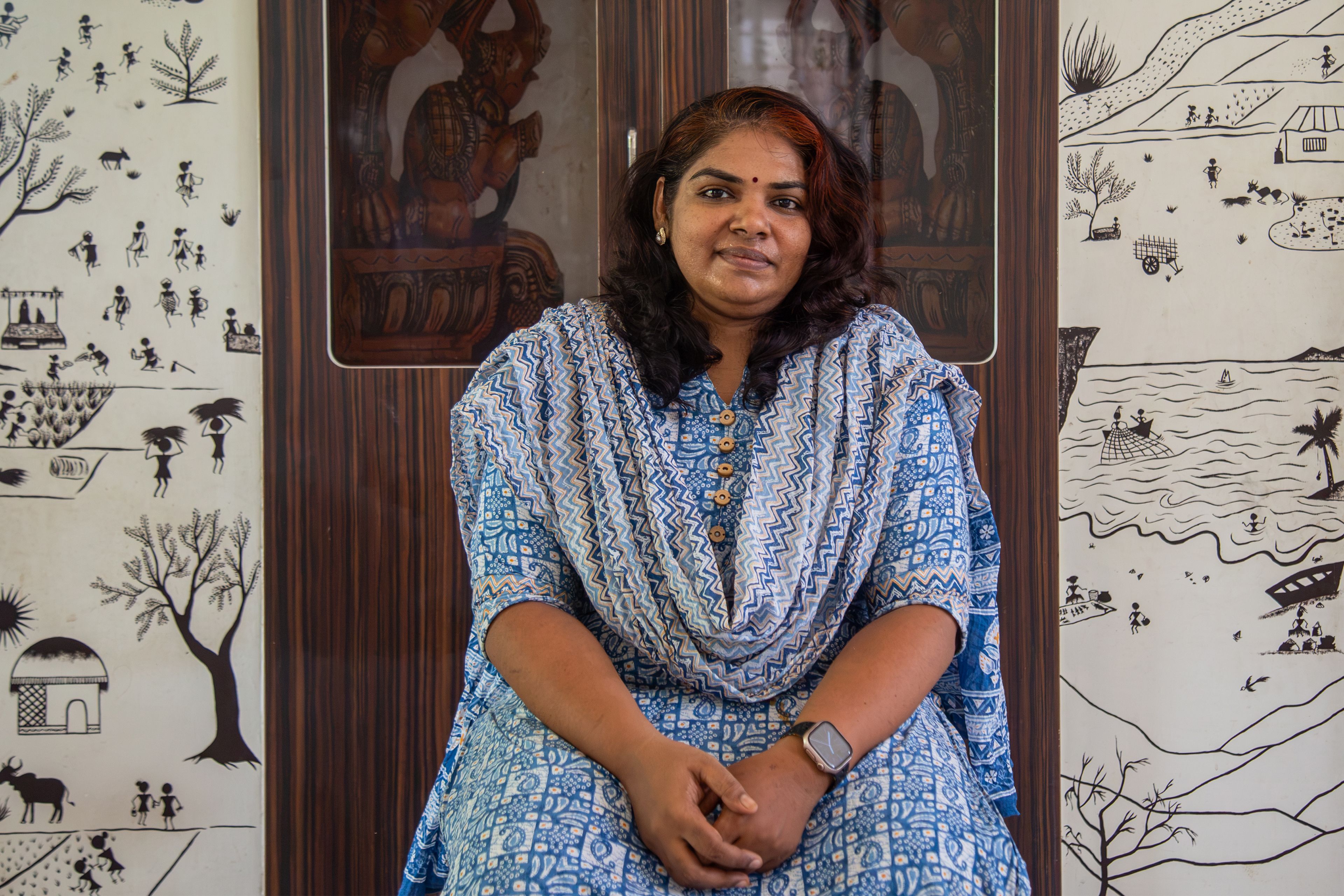 A Tamil woman wearing a blue patterned dress and scarf sits in front of wood-pannelled doors and walls papered with simple illustrations portraying an island community and life. The woman has her nose pierced and brown-red hair that finishes just below her shoulders. She is smiling.