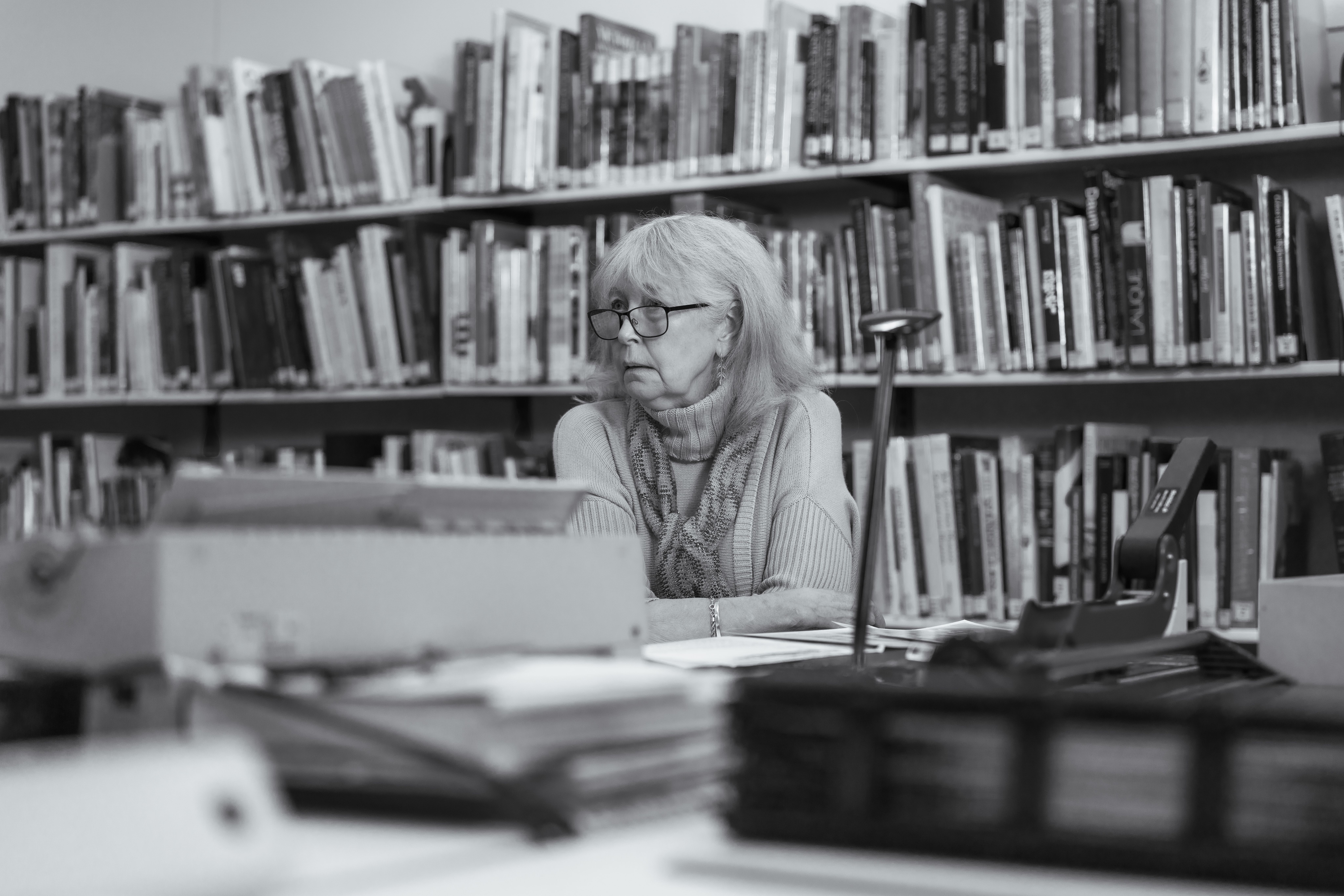 Black and white portrait of a woman (Denise Larcombe) looking to her right as she leans on a table scattered with documents and objects, the library shelf in the background.