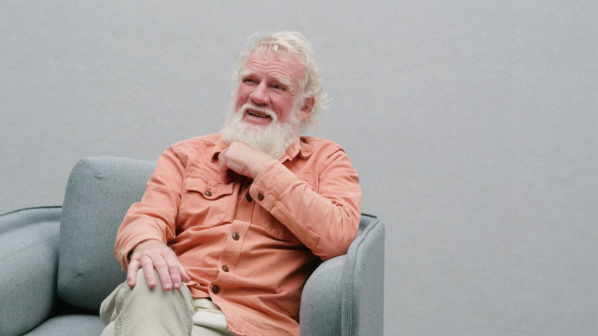 Bruce Pascoe sits in a grey arm chair wearing a peach coloured button up shirt, he has a white beard and is looking upwards and towards the right.