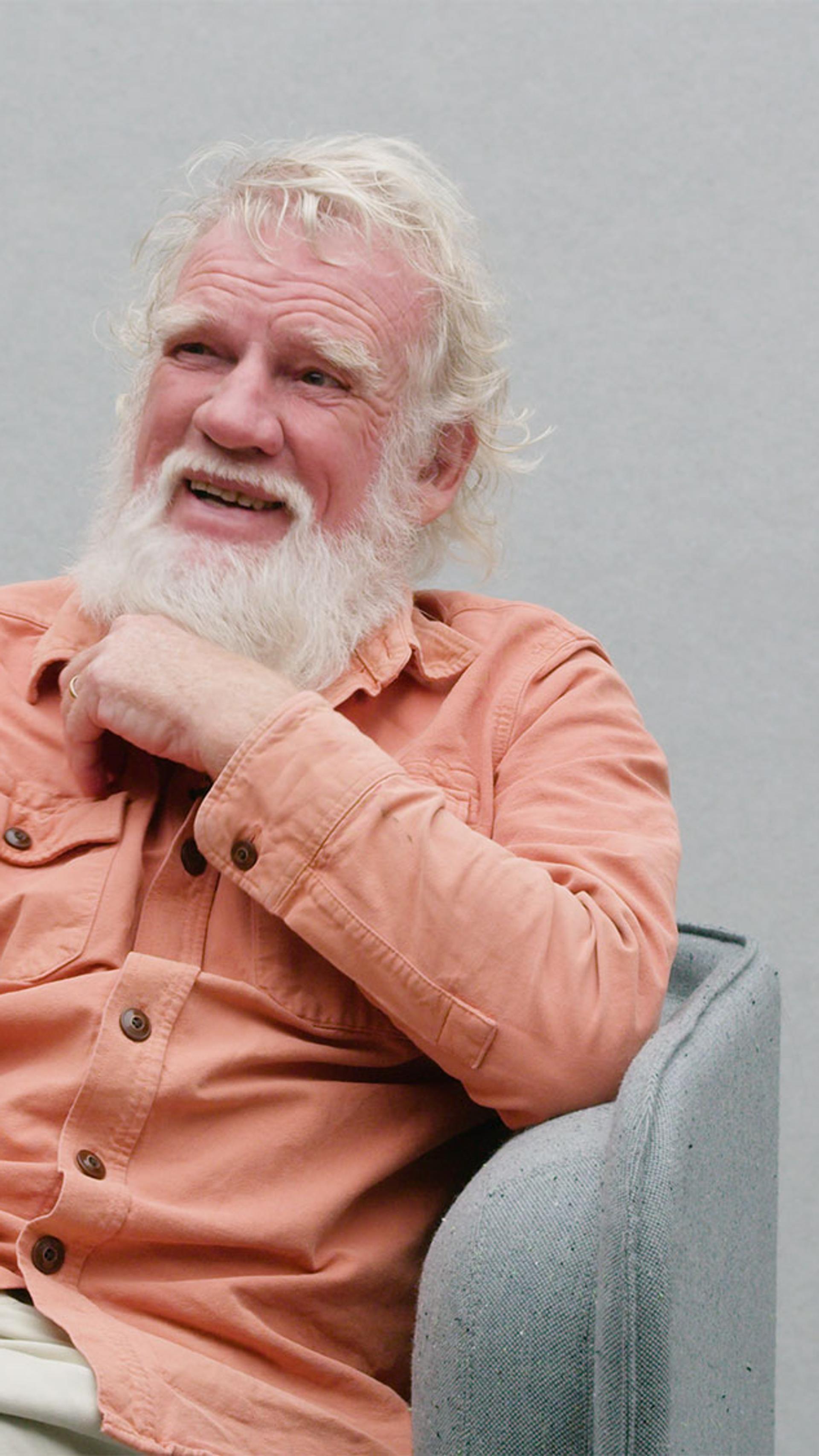 Bruce Pascoe sits in a grey arm chair wearing a peach coloured button up shirt, he has a white beard and is looking upwards and towards the right.