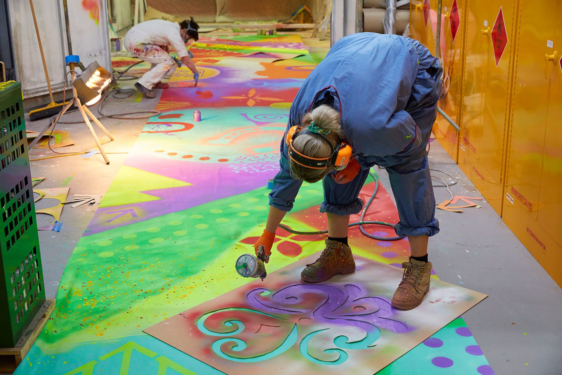 A figure leans over with an airbrush painting a stencilled shape in purple onto a blue surface.