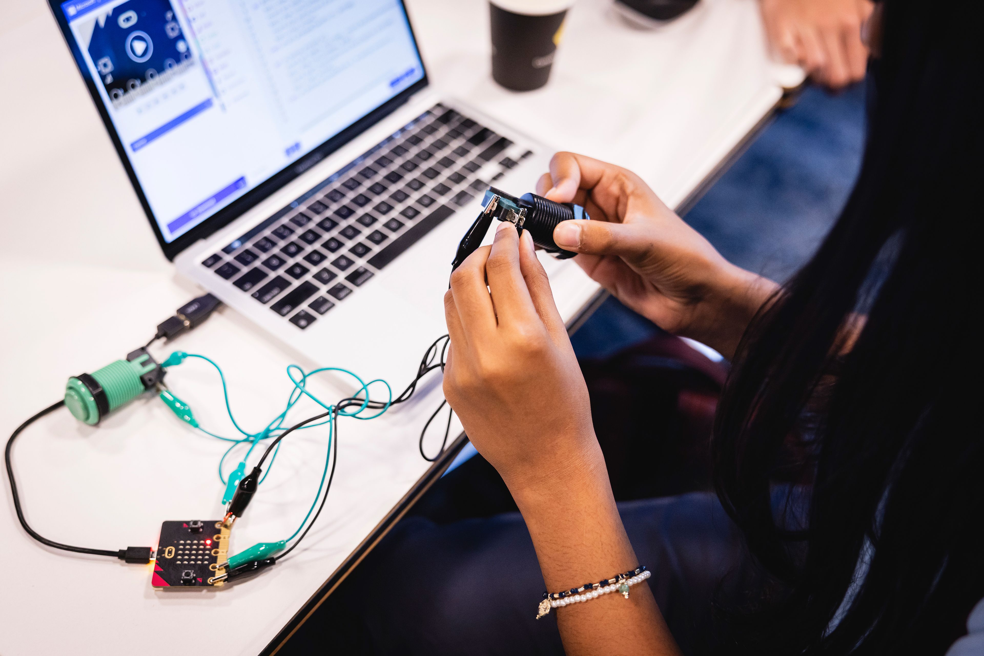 Close-up photo over a student’s shoulder holding a mechanical part with wires. The wires connect to a laptop on a table.