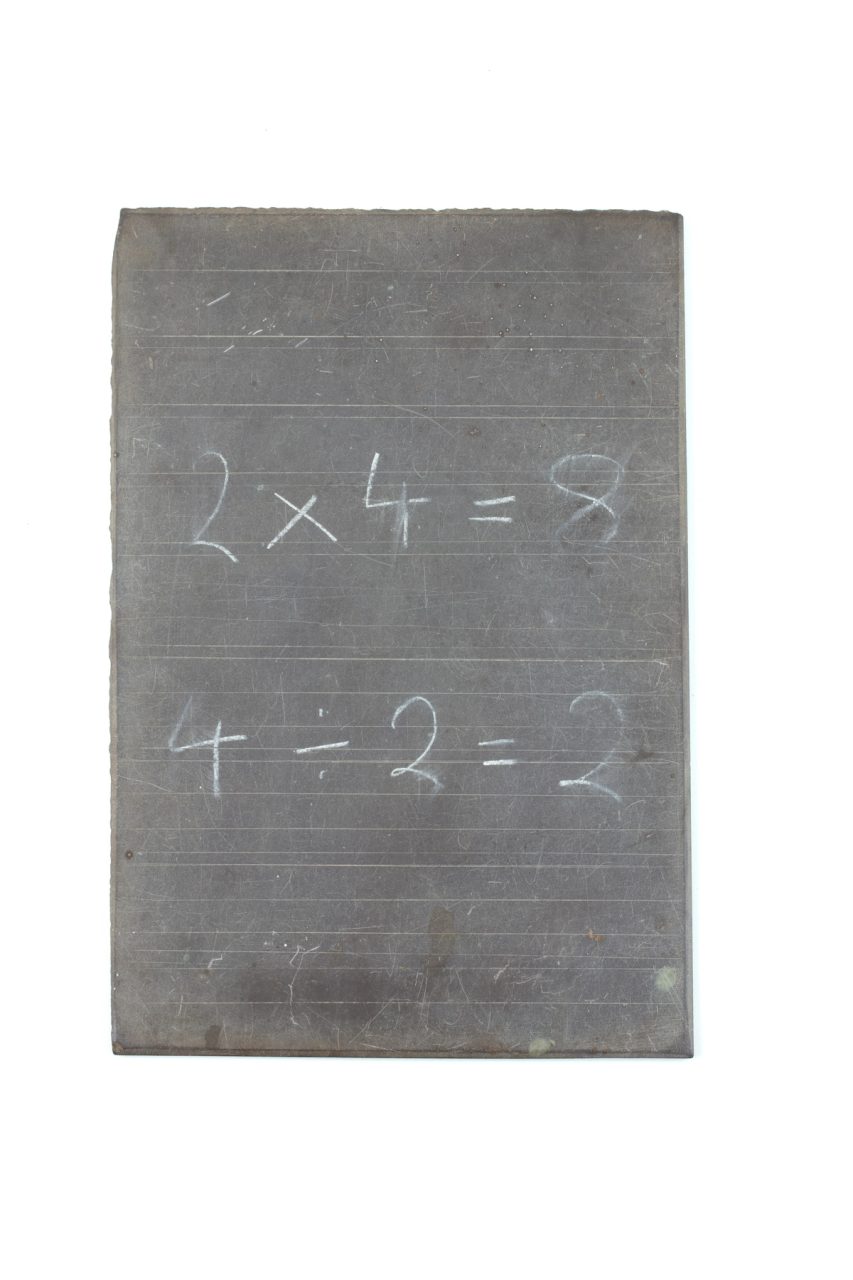 An old school slate, with fine ruled guide lines and some math calculations written on white chalk, '2x4=8, 4÷2=2'.