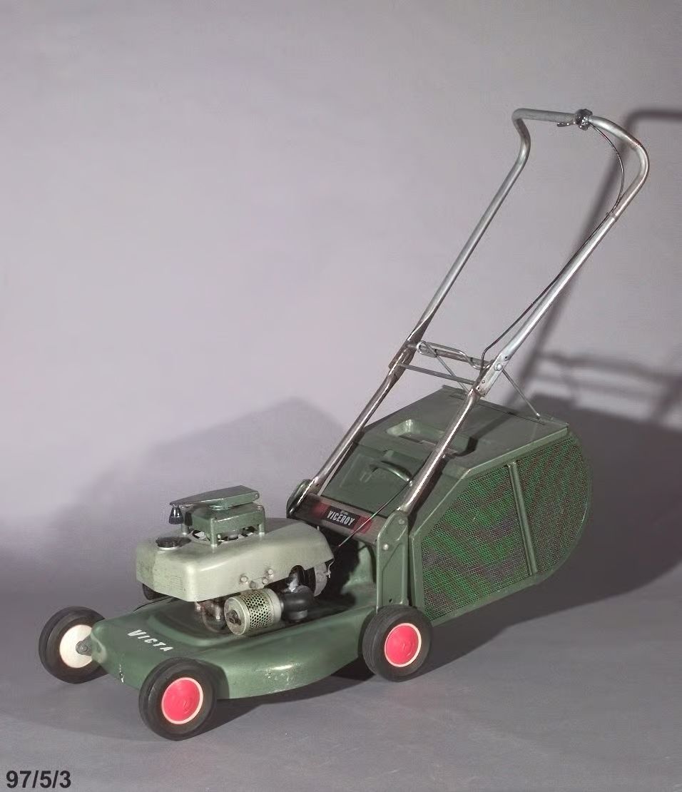 Photo of a two tone green hammertone lawnmower, painted metal base with four plastic and rubber wheels on concealed axles. The base supports a vertically mounted two stroke engine covered by a metal petrol tank and cowl.