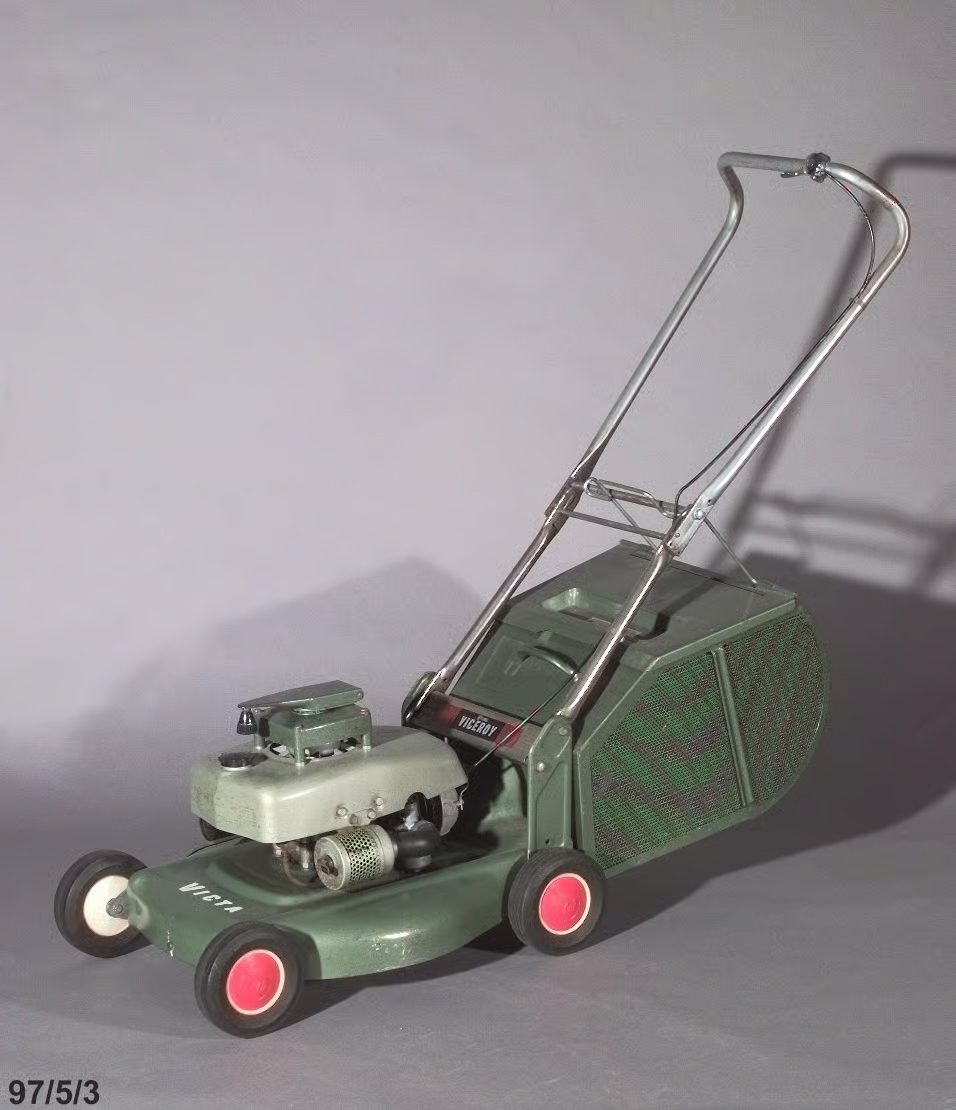 Photo of a two tone green hammertone lawnmower, painted metal base with four plastic and rubber wheels on concealed axles. The base supports a vertically mounted two stroke engine covered by a metal petrol tank and cowl.
