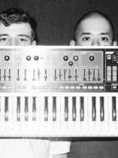Two fellas hold a single keyboard synth in front of their faces