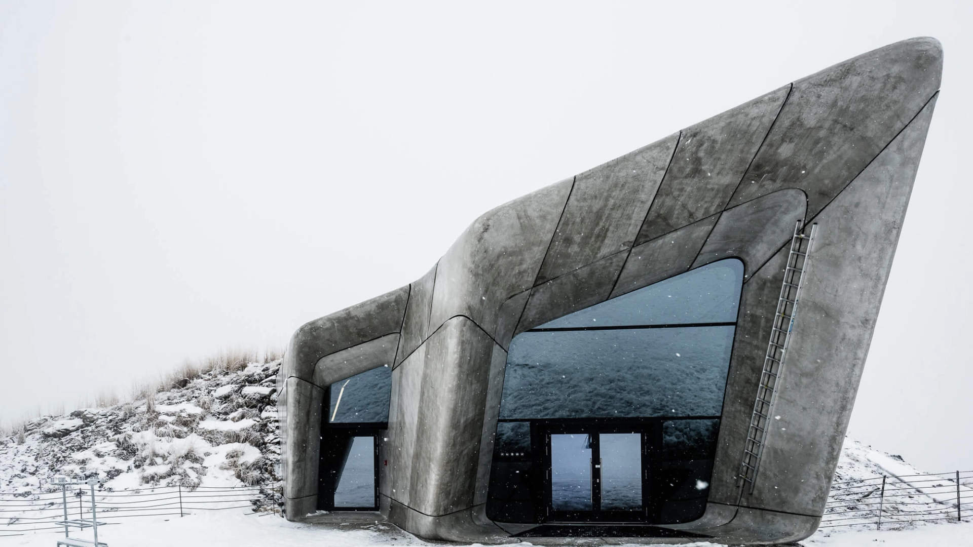 Brutalist grey structures with large windows appear out of a mound in the snowy ground, under a white sky.