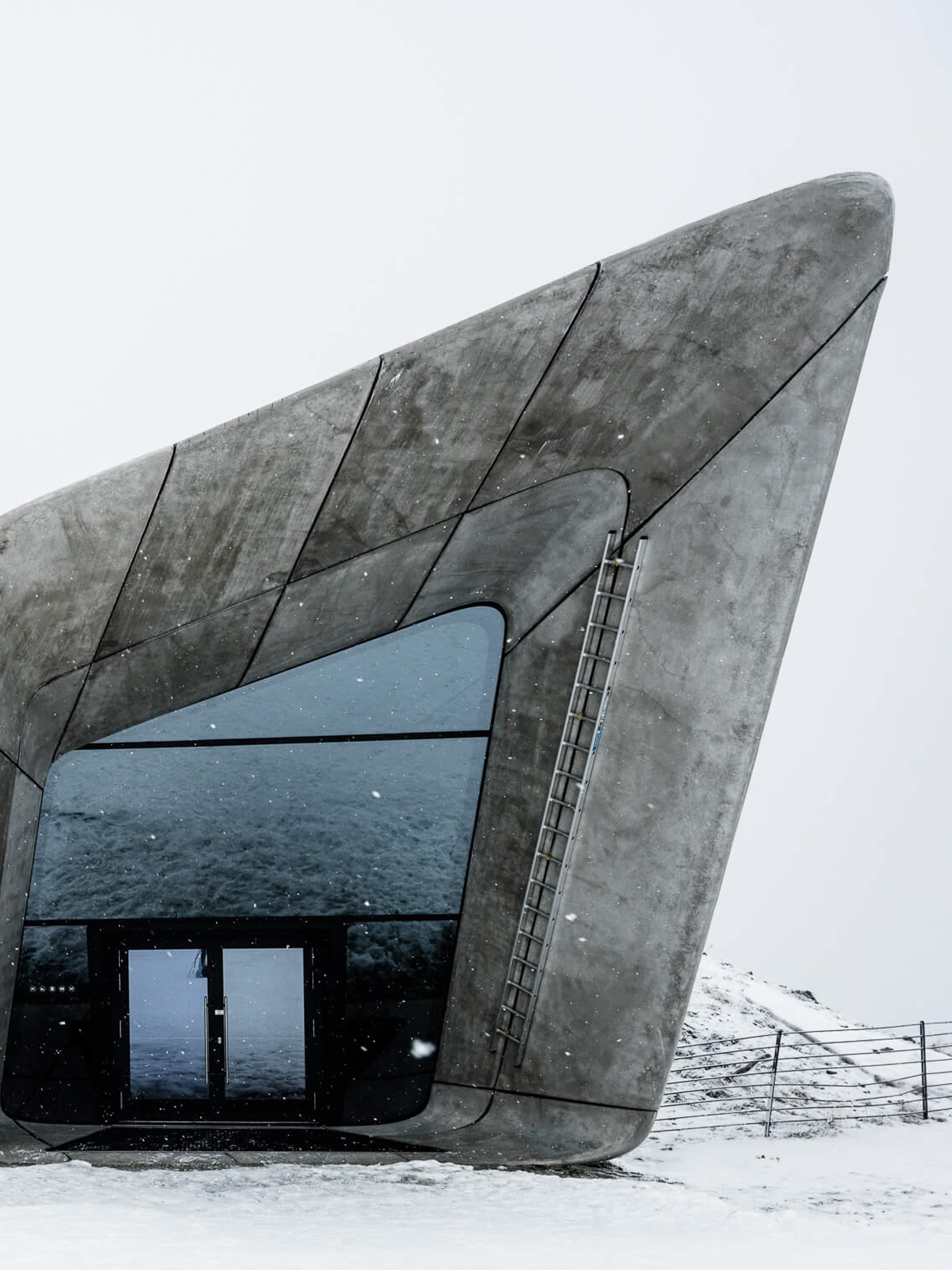 Brutalist grey structures with large windows appear out of a mound in the snowy ground, under a white sky.