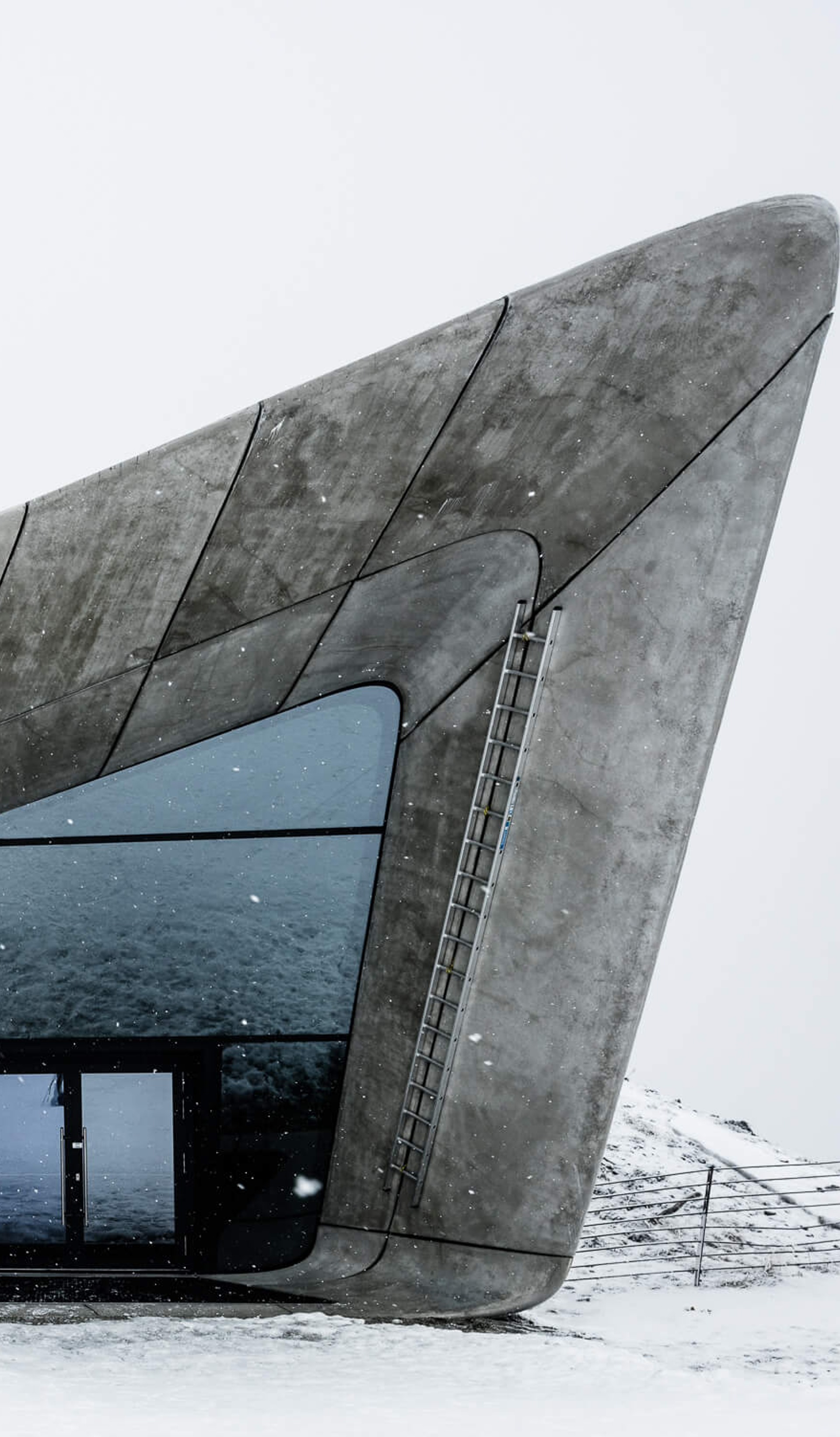 Brutalist grey structures with large windows appear out of a mound in the snowy ground, under a white sky.