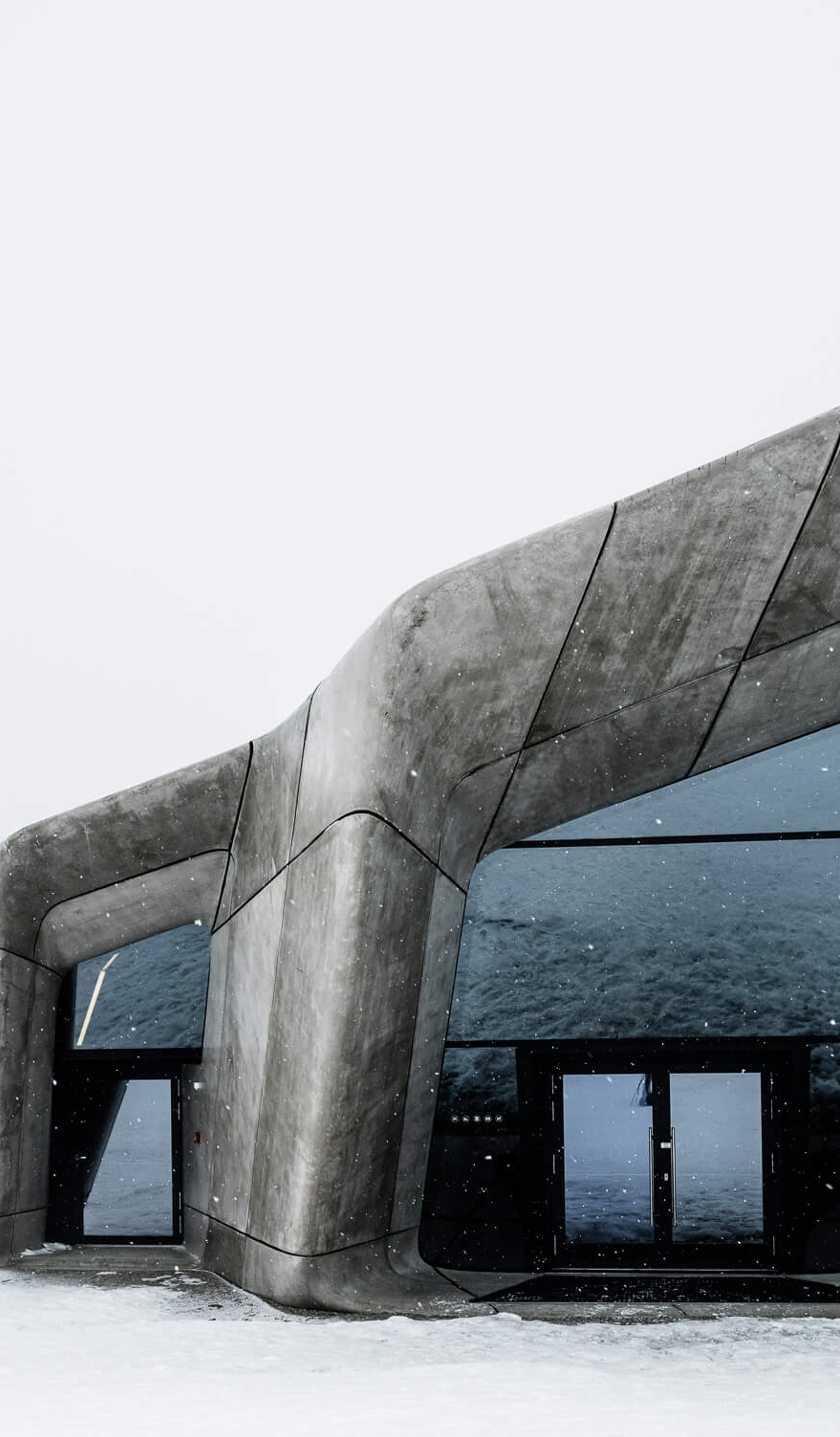 Brutalist grey structures with large windows appear out of a mound in the snowy ground, under a white sky.