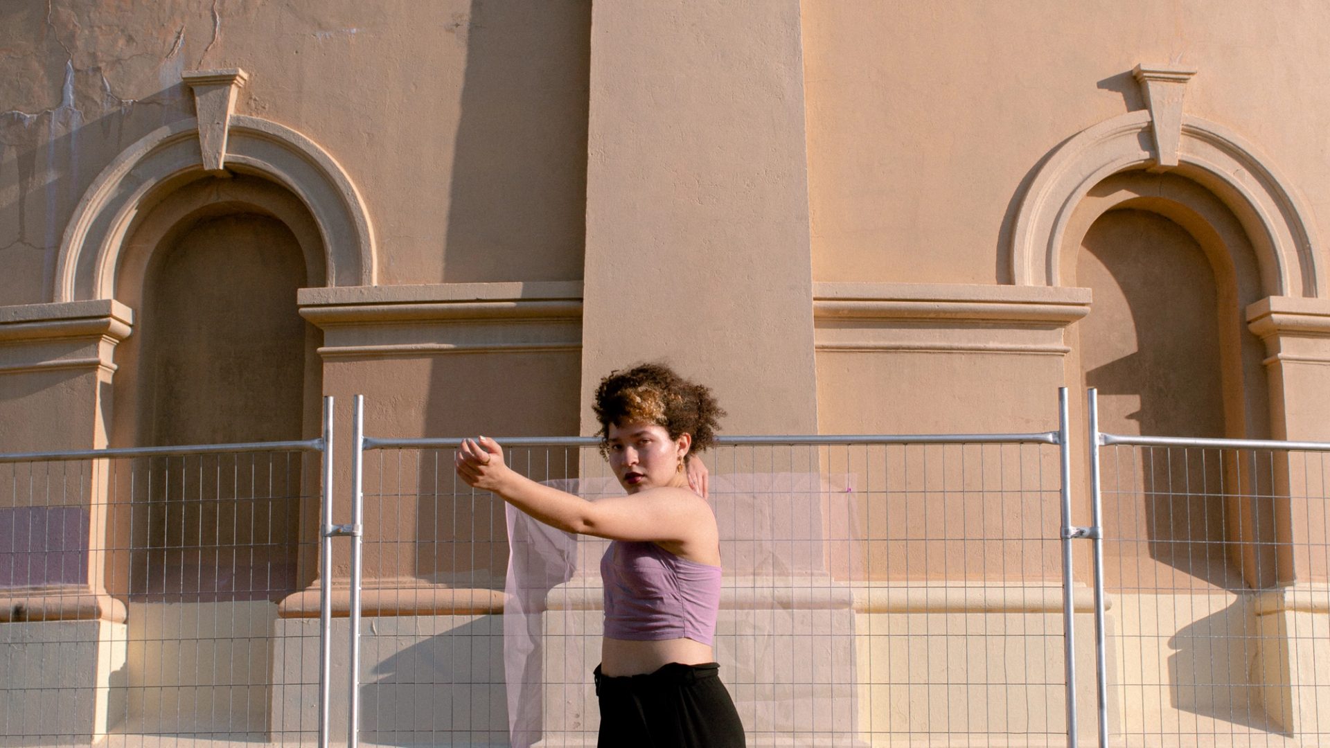 A woman in a purple top black pants and white shoes stands on the tips of her toes in front of a fenced-off beige building.
