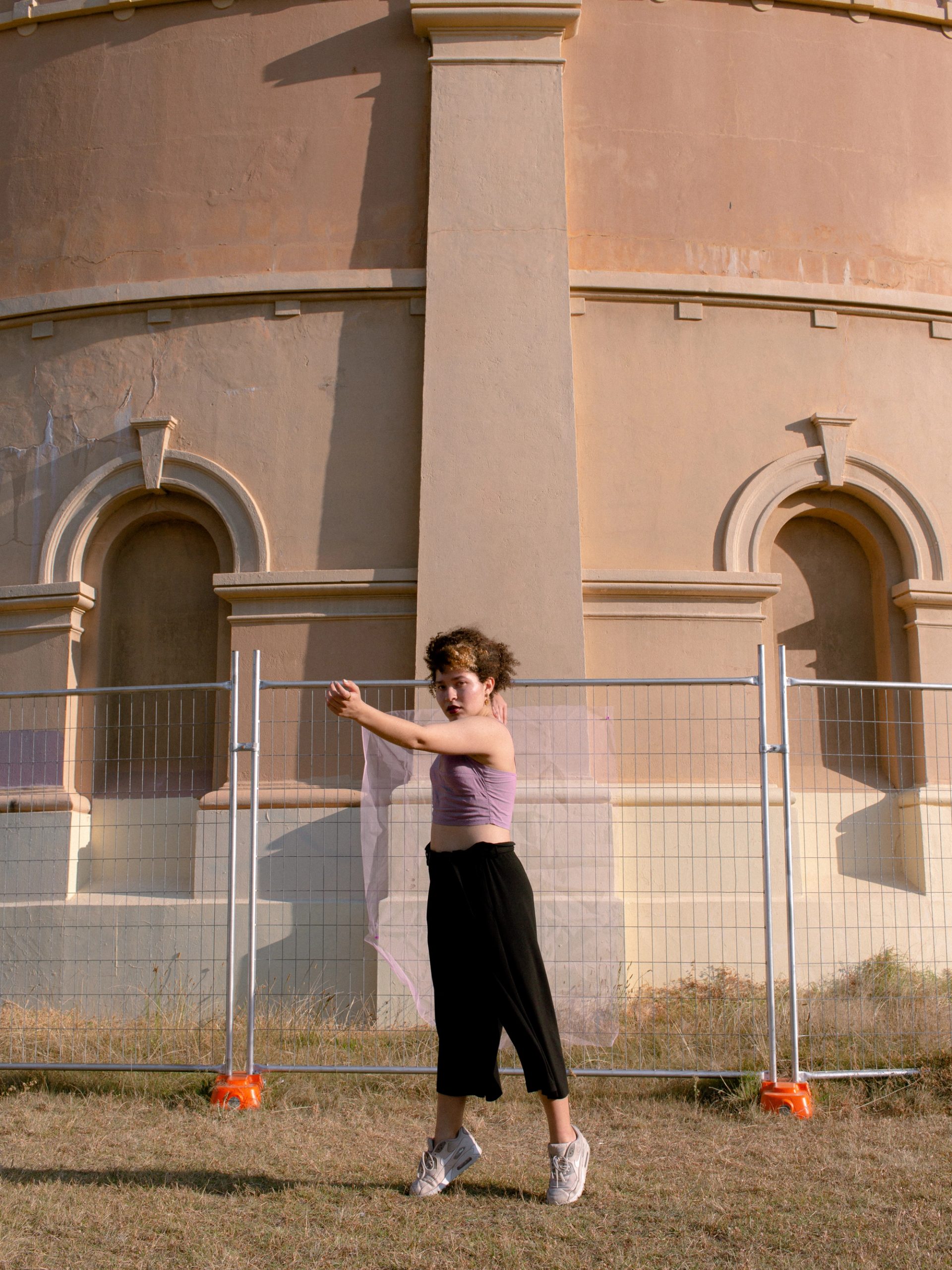 A woman in a purple top black pants and white shoes stands on the tips of her toes in front of a fenced-off beige building.
