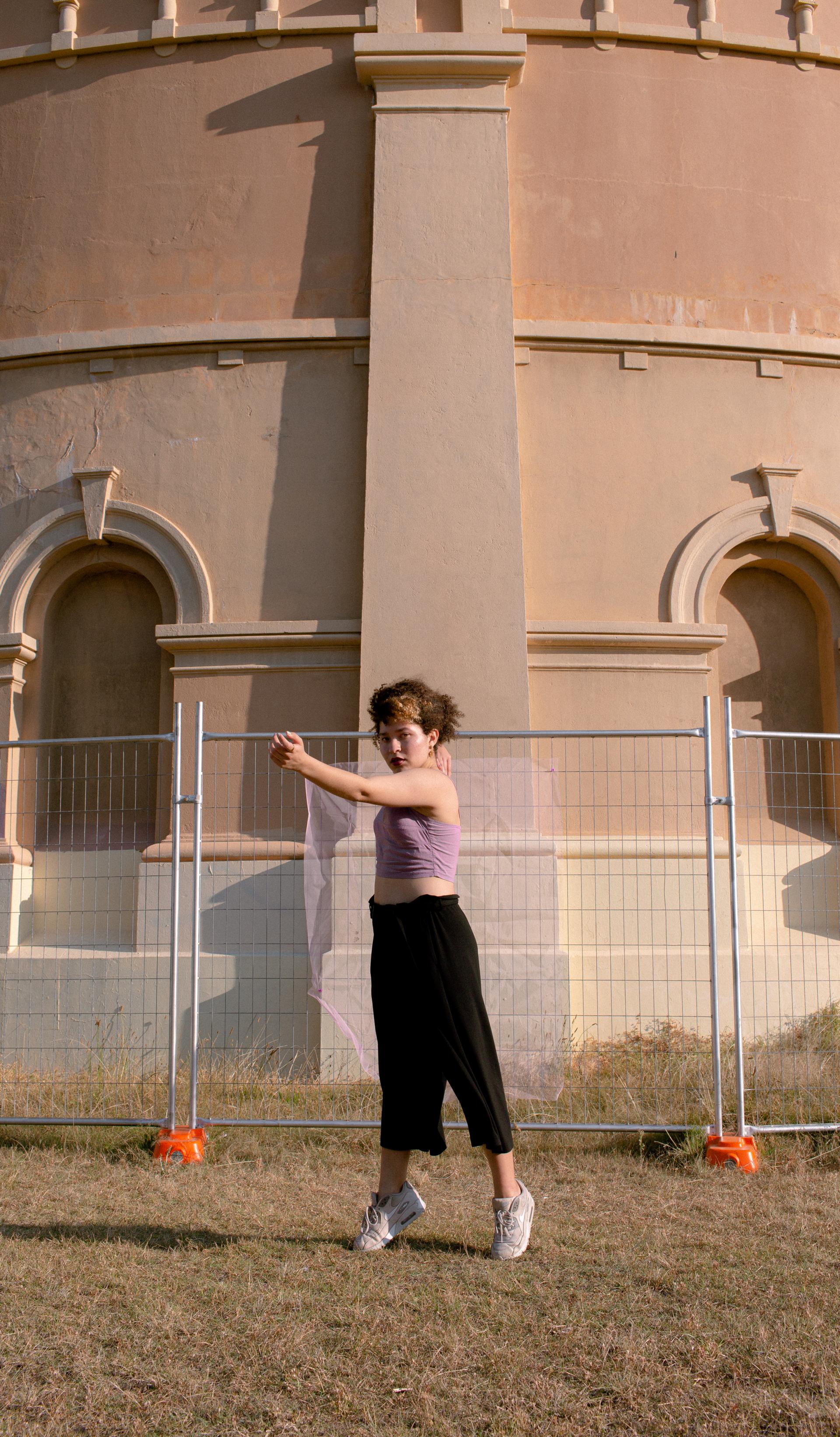 A woman in a purple top black pants and white shoes stands on the tips of her toes in front of a fenced-off beige building.