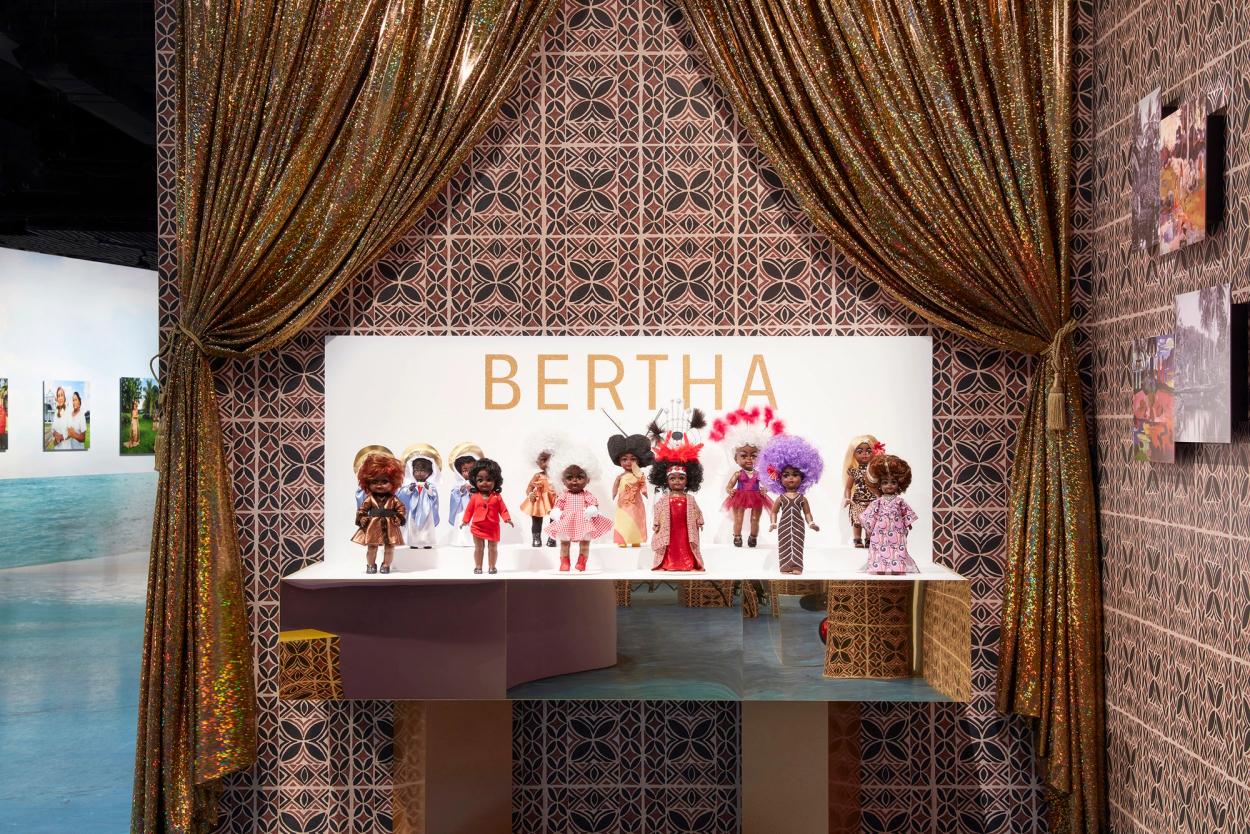 13 small plastic brown female dolls, each dressed in different outfits, stand on a small white platform.