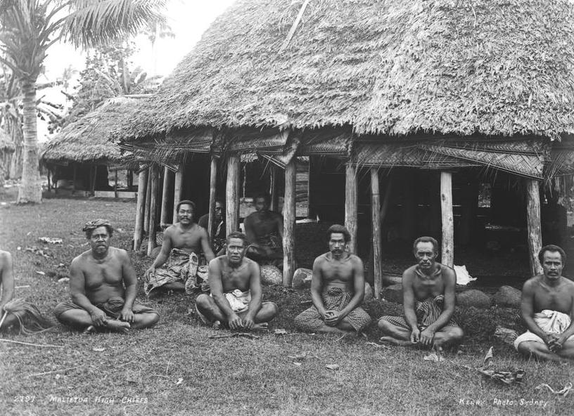 a group of men sitting cross legged in front of and under a traditional Samoan dwelling