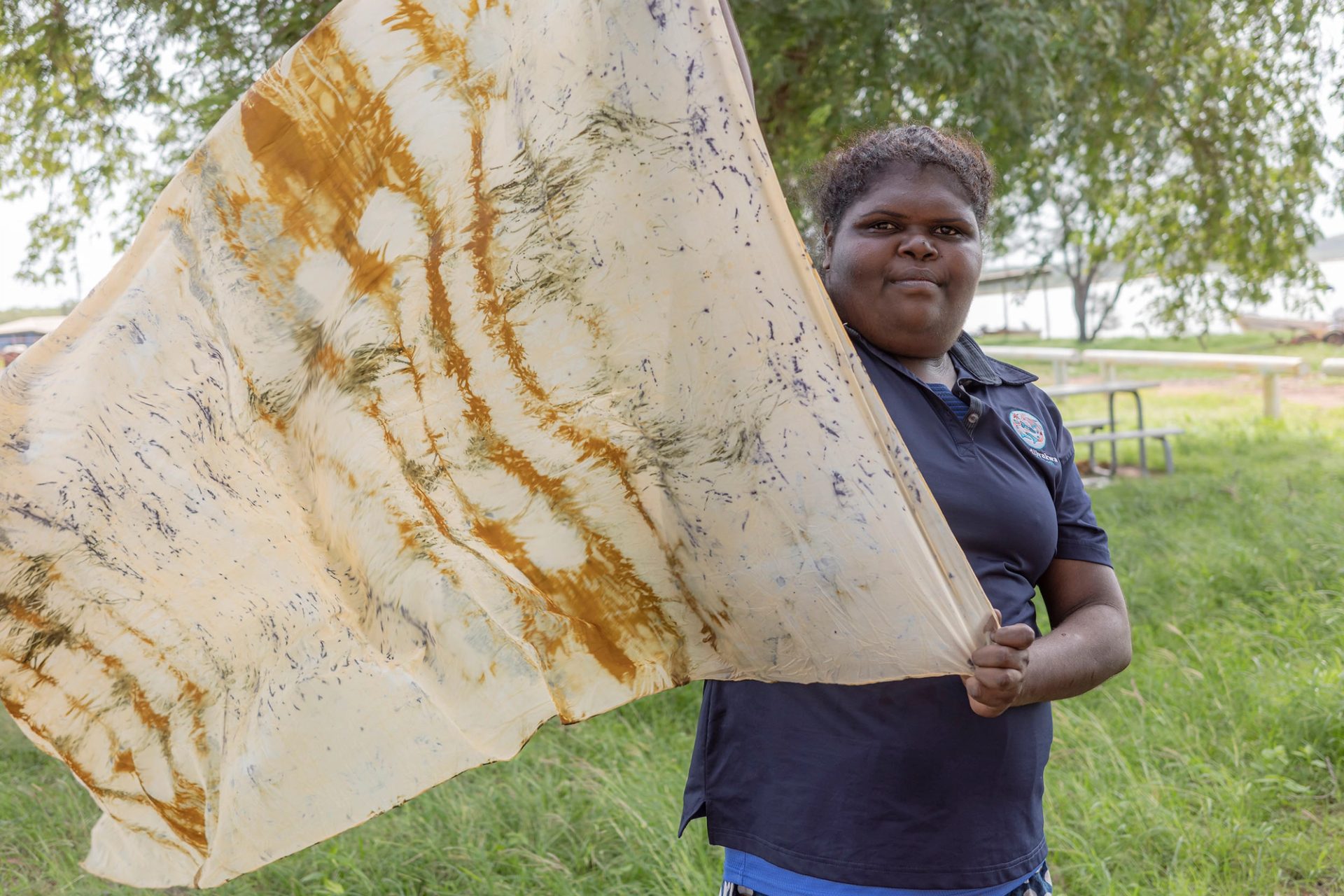 A woman stands outside holding a dyed piece of silk textile as it blows in the wind.