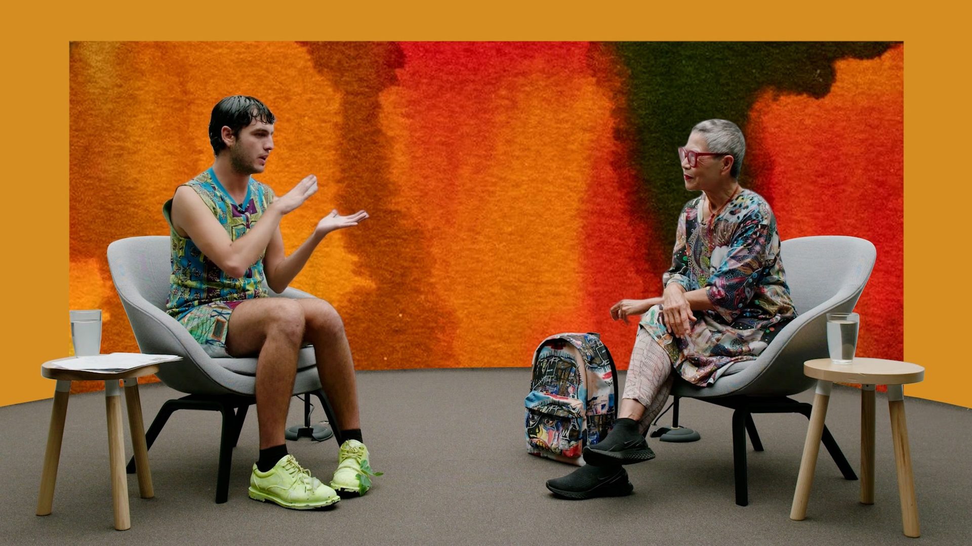 Jordan Gogos and Jenny Kee sit facing each other on grey chairs with an orange painted background.