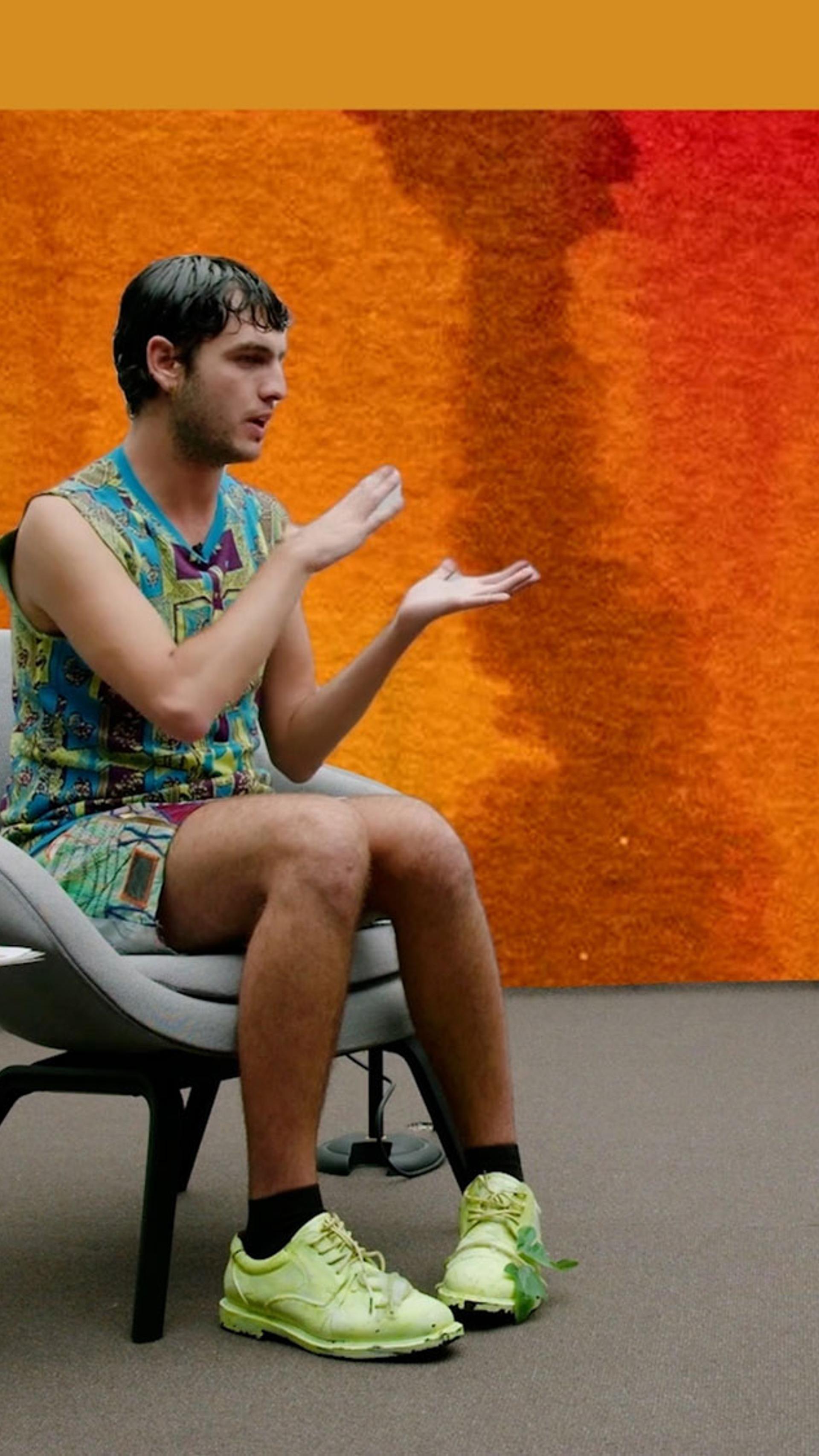 Jordan Gogos and Jenny Kee sit facing each other on grey chairs with an orange painted background.