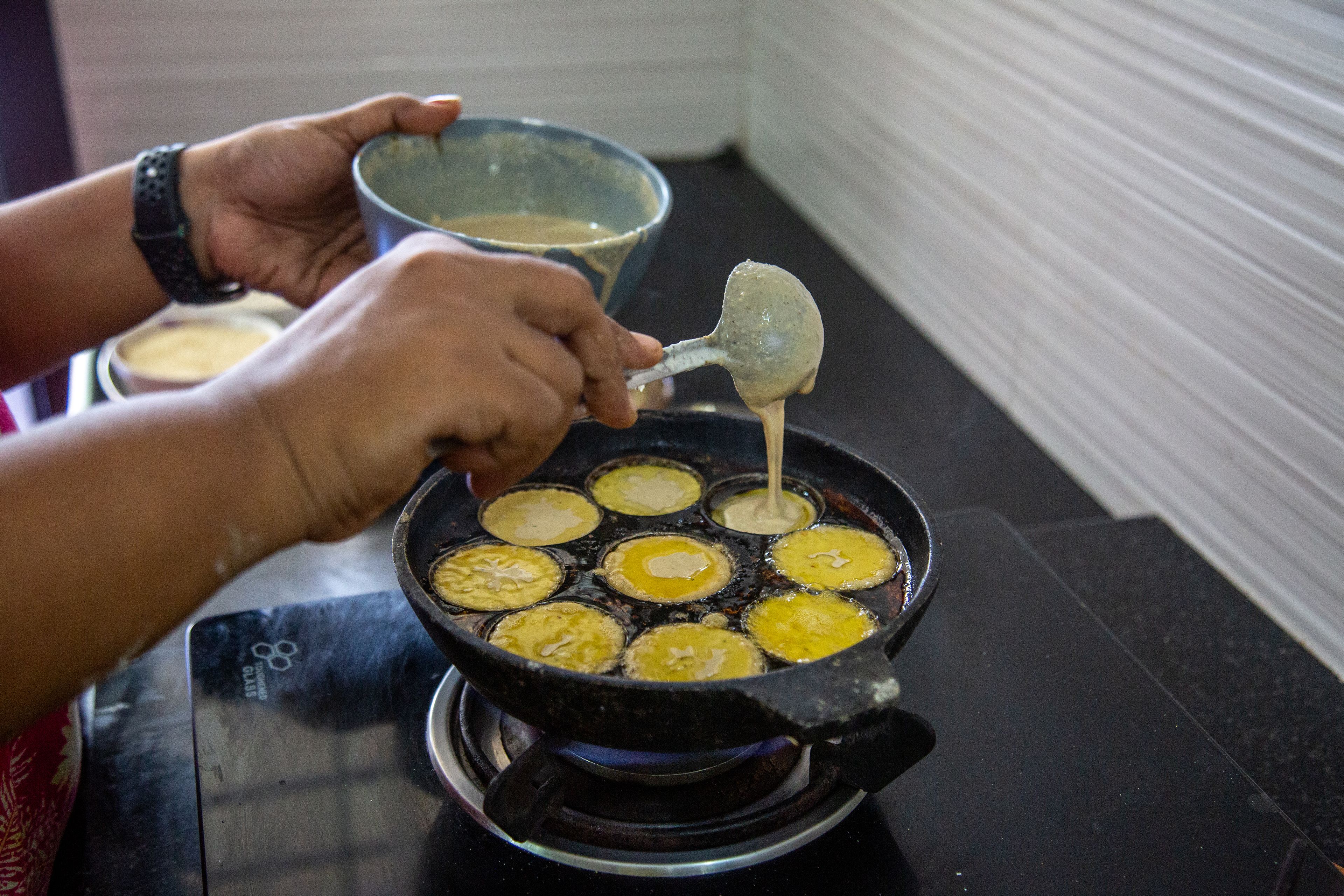 Brown hands ladle batter into a paniyaram pan.