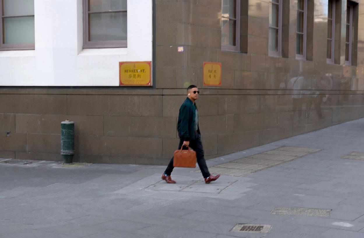 A figure in dark sunglasses, a black jacket and carrying a brown suitcase walks on the footpath.