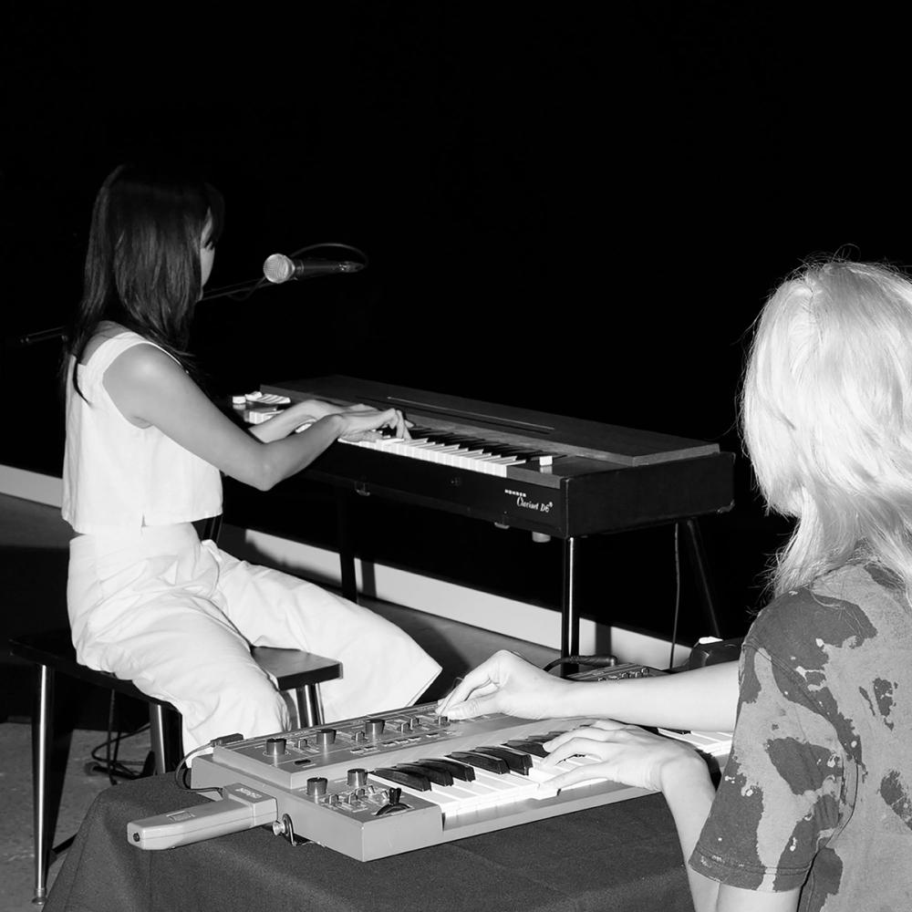 Black and white image of two people, both seated at keyboards, seen from behind.