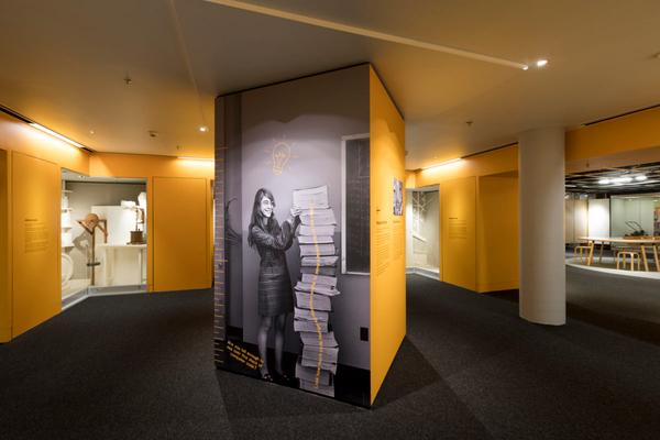 A yellow wall with a life size illustration of Margaret Hamilton, standing beside a long list of code.