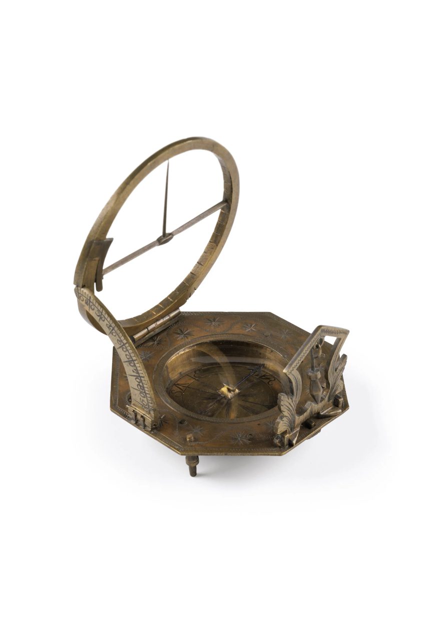 A sundial compass against a white background: made of brass, it is an intricate machine, with a circular folded out section bisected by the compass.