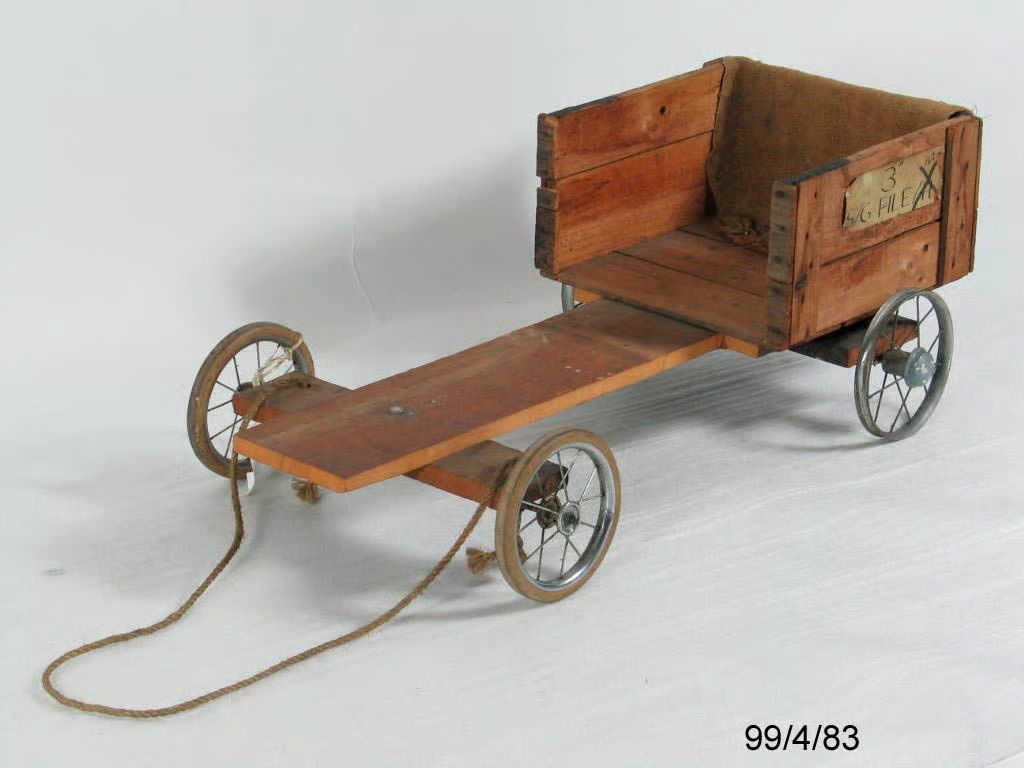 Billycart. The chassis of this billycart is made from wood. A plank forms the central axis. A wooden crate, cut down to four sides, forms the cabin and seat. A brown hessian bag has been draped over the back of this crate. Stuck on the left side of the crate is a piece of paper with the inscription; '3" 162 S/G FILE/H'.