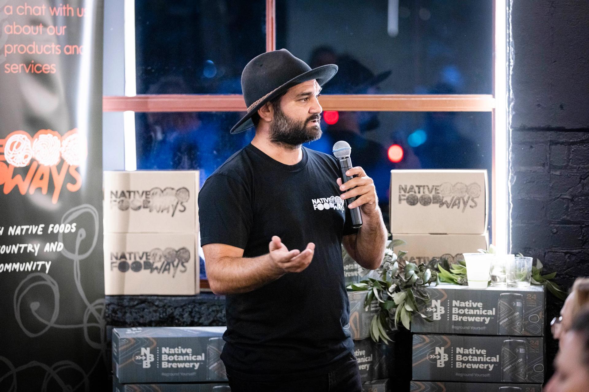 Man speaks on microphone to crowd, with boxes of Native Foodways and Native Botanical Brewery around them